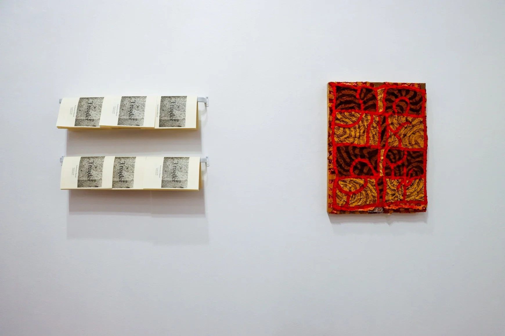 A photograph that shows several copies of a black-and-white xeroxed and stapled instruction manual hanging from horizontal shelves mounted to a white wall. Next to them hangs a painting in muddy hues of orange, yellow, and red.