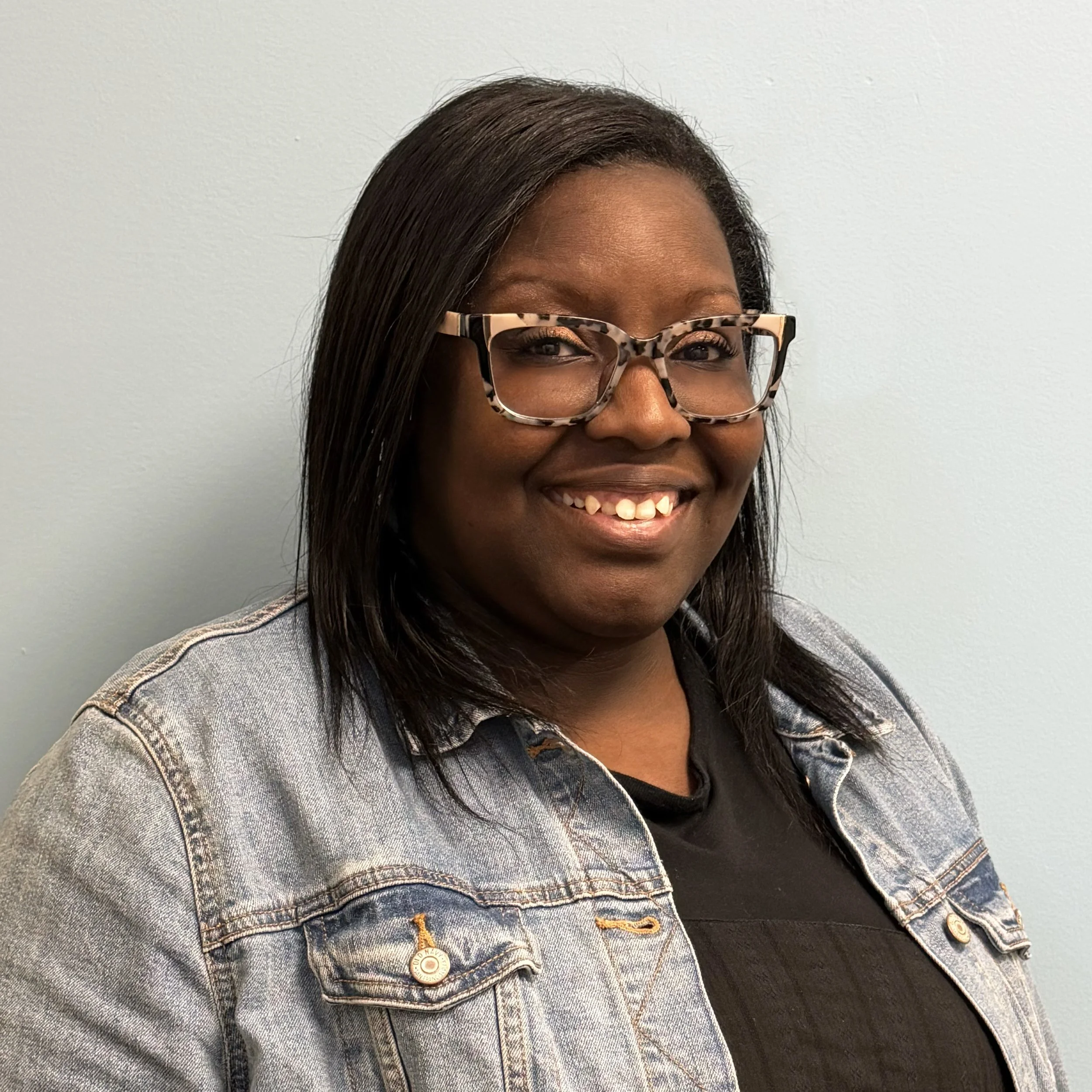 A woman with dark hair, glasses with tortoise shell frames, smiling, wearing a denim jacket over a black shirt, standing against a light-colored wall.