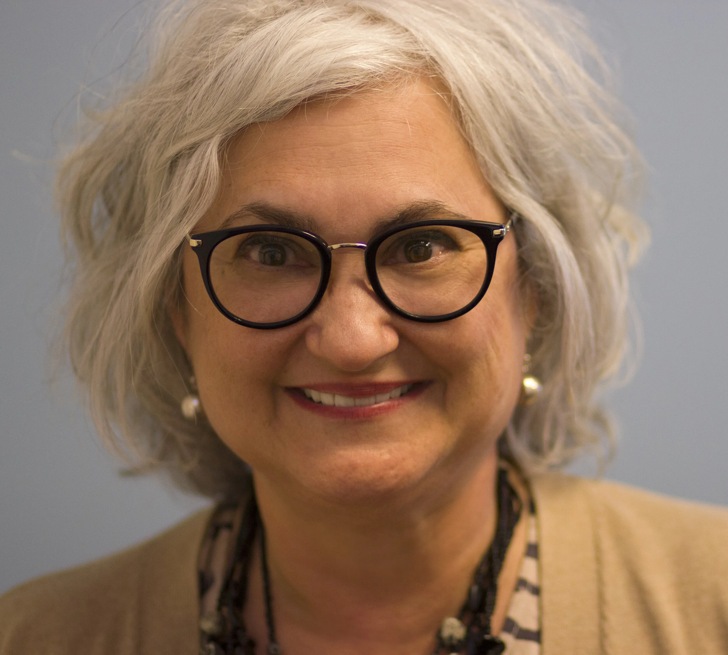 A smiling woman with curly gray hair wearing black glasses, pearl earrings, a beige blazer, and a patterned top, standing in front of a light blue background.