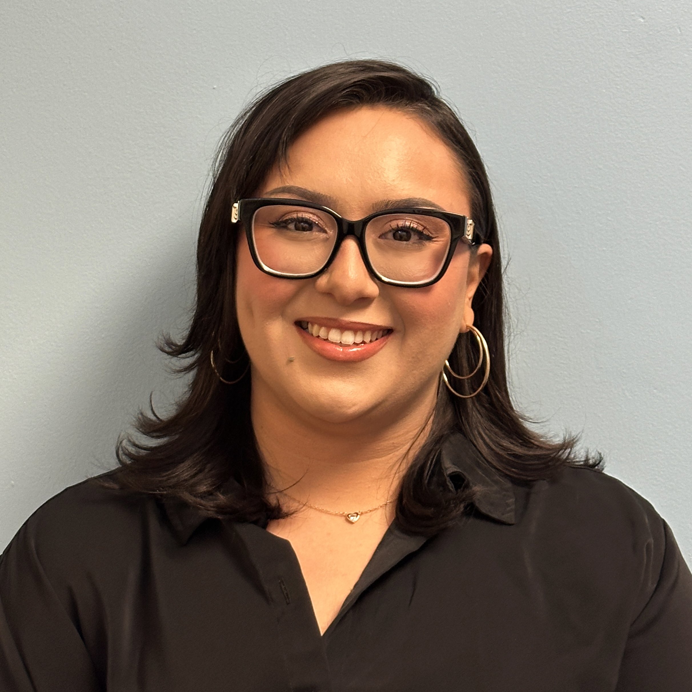 A smiling woman with dark hair, glasses, hoop earrings, wearing a black shirt, standing against a light blue wall.