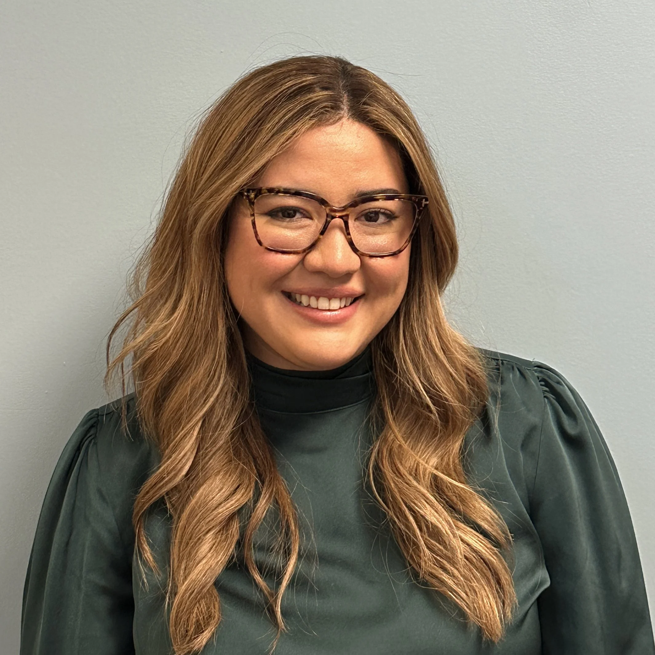 Smiling woman with long wavy hair, wearing glasses and a dark green top, standing against a light-colored wall.