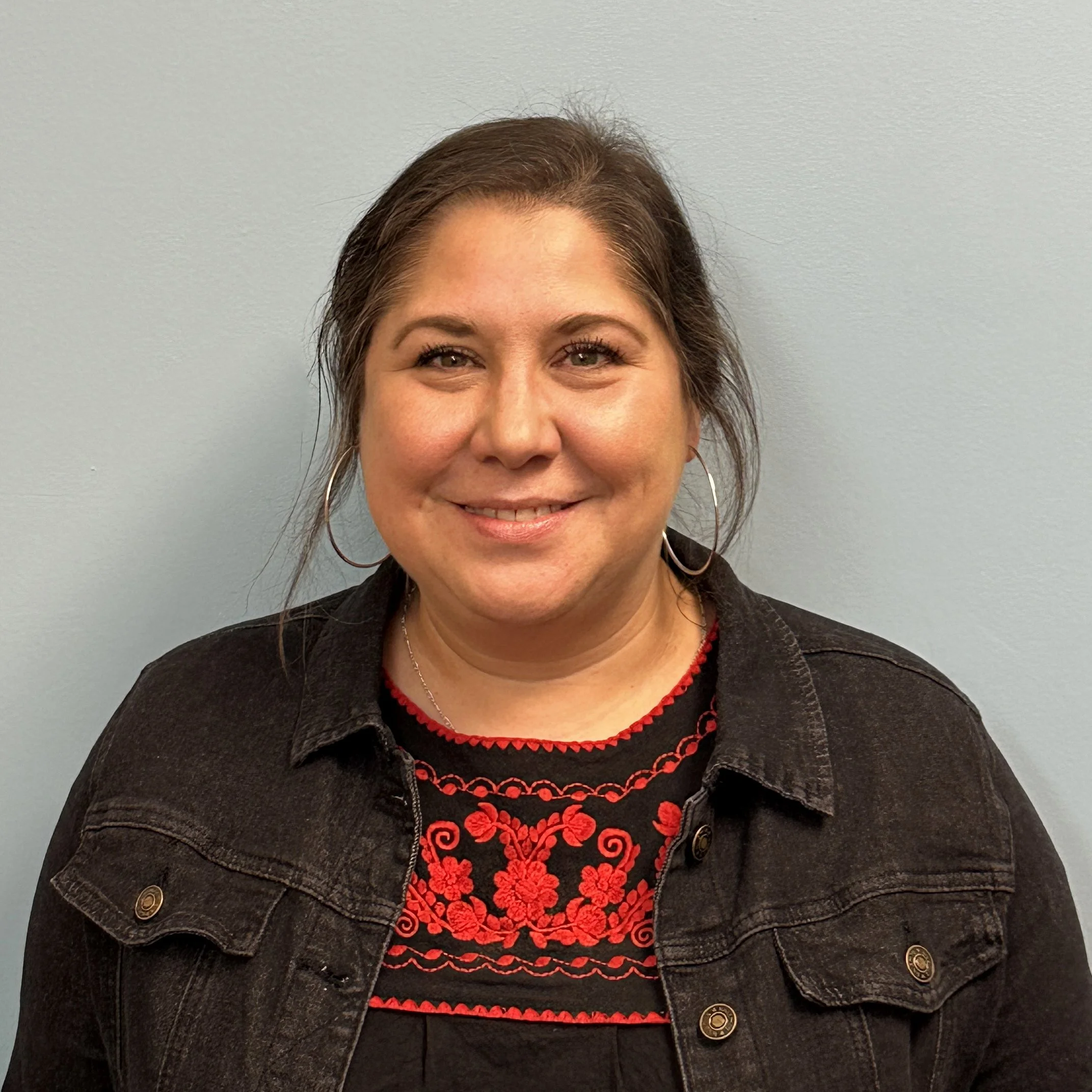 A smiling woman with light skin, shoulder-length brown hair, wearing hoop earrings, a black embroidered top with red floral design, and a black denim jacket, standing against a plain light blue wall.