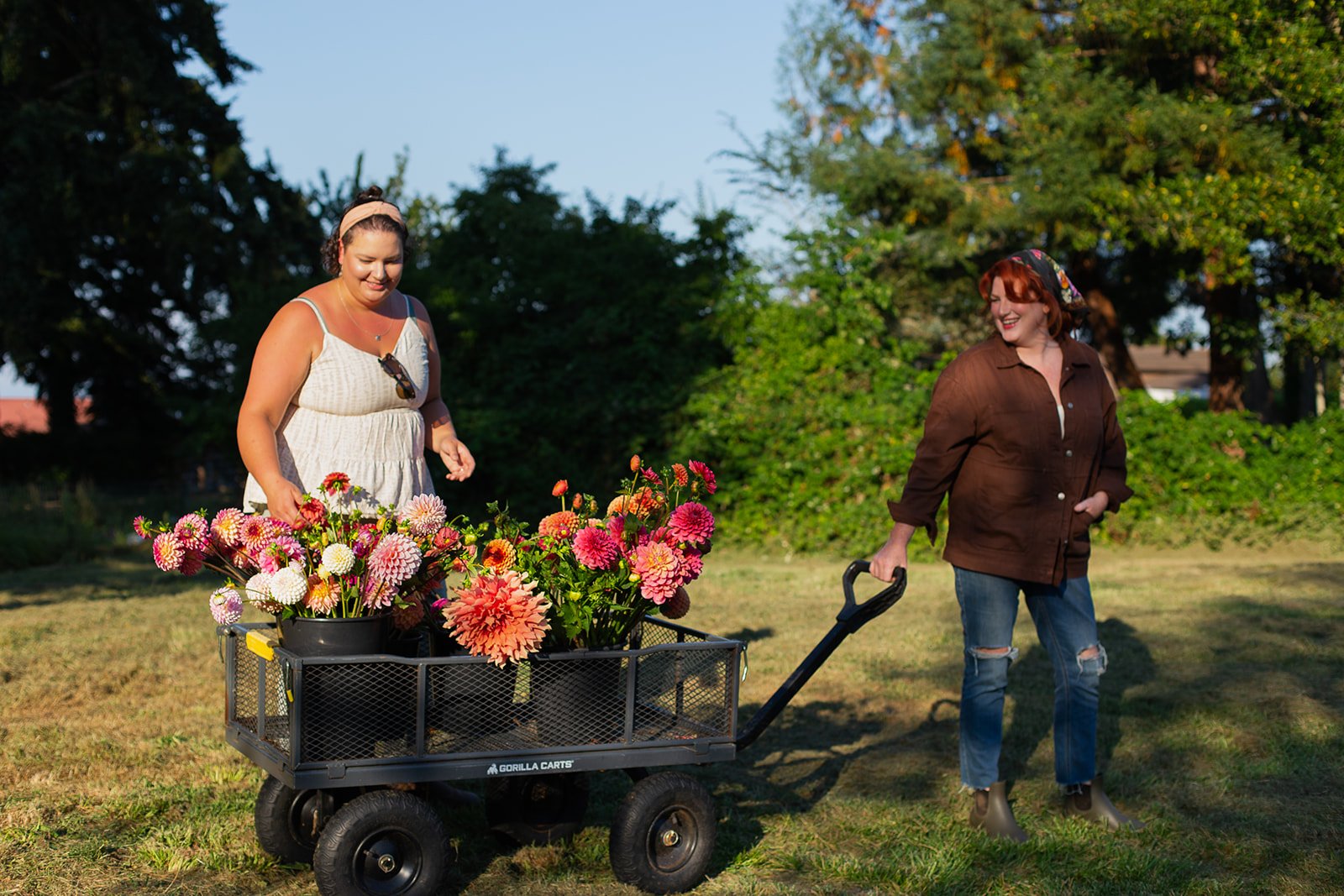 Two women smiling outdoors with one pushing a cart filled with colorful flowers on a grassy area with trees in the background.