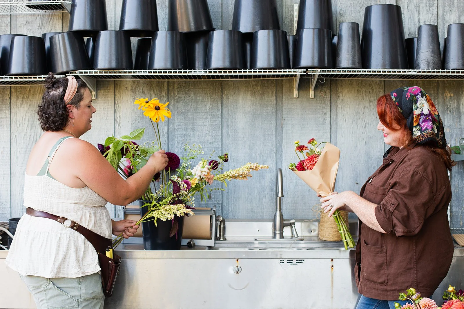 Two women exchanging flowers in a flower shop or market, one handing a bouquet to the other, with shelves of black pots above them and a sink in the background.