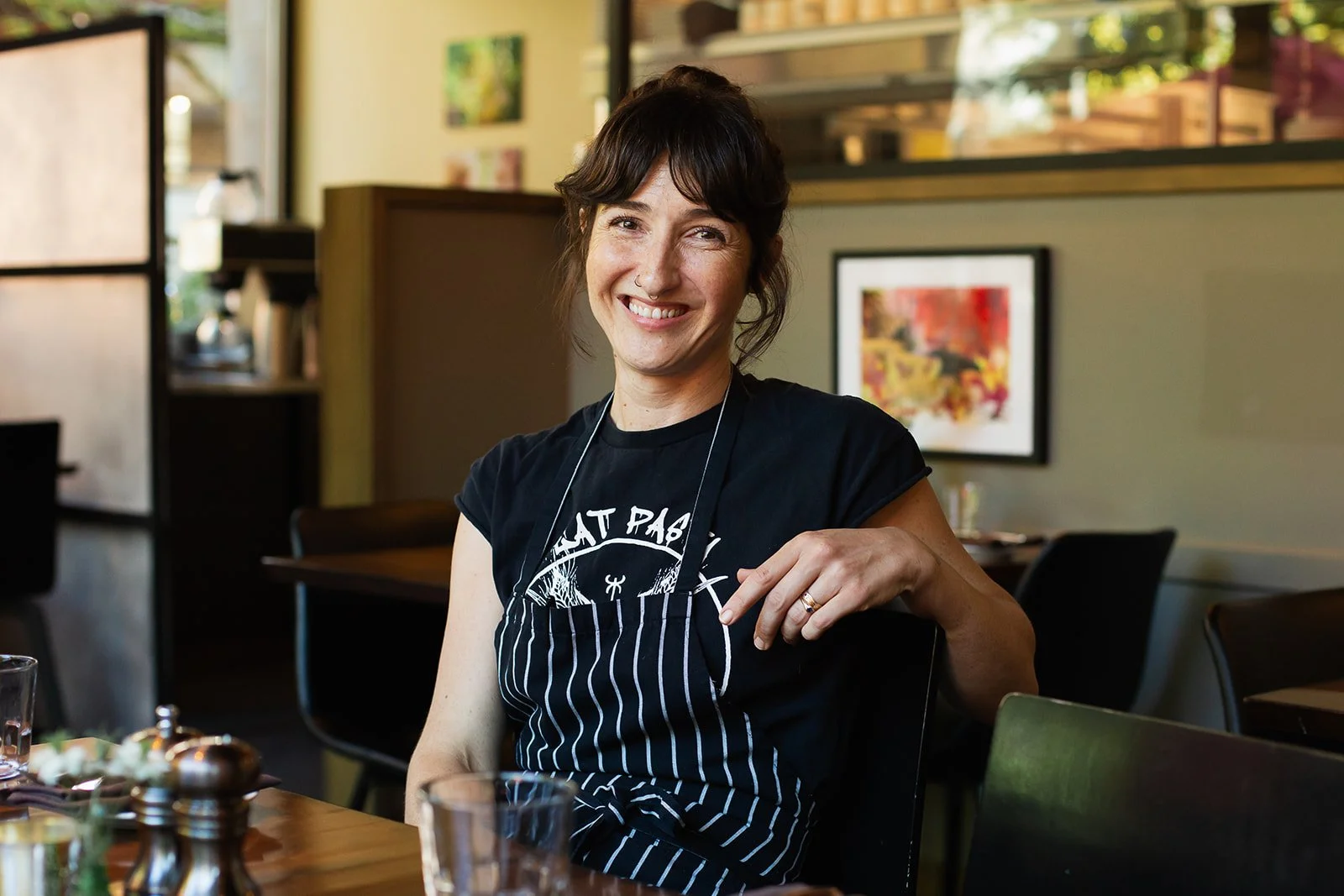 A woman with short dark hair and a striped apron, smiling while sitting at a restaurant table.