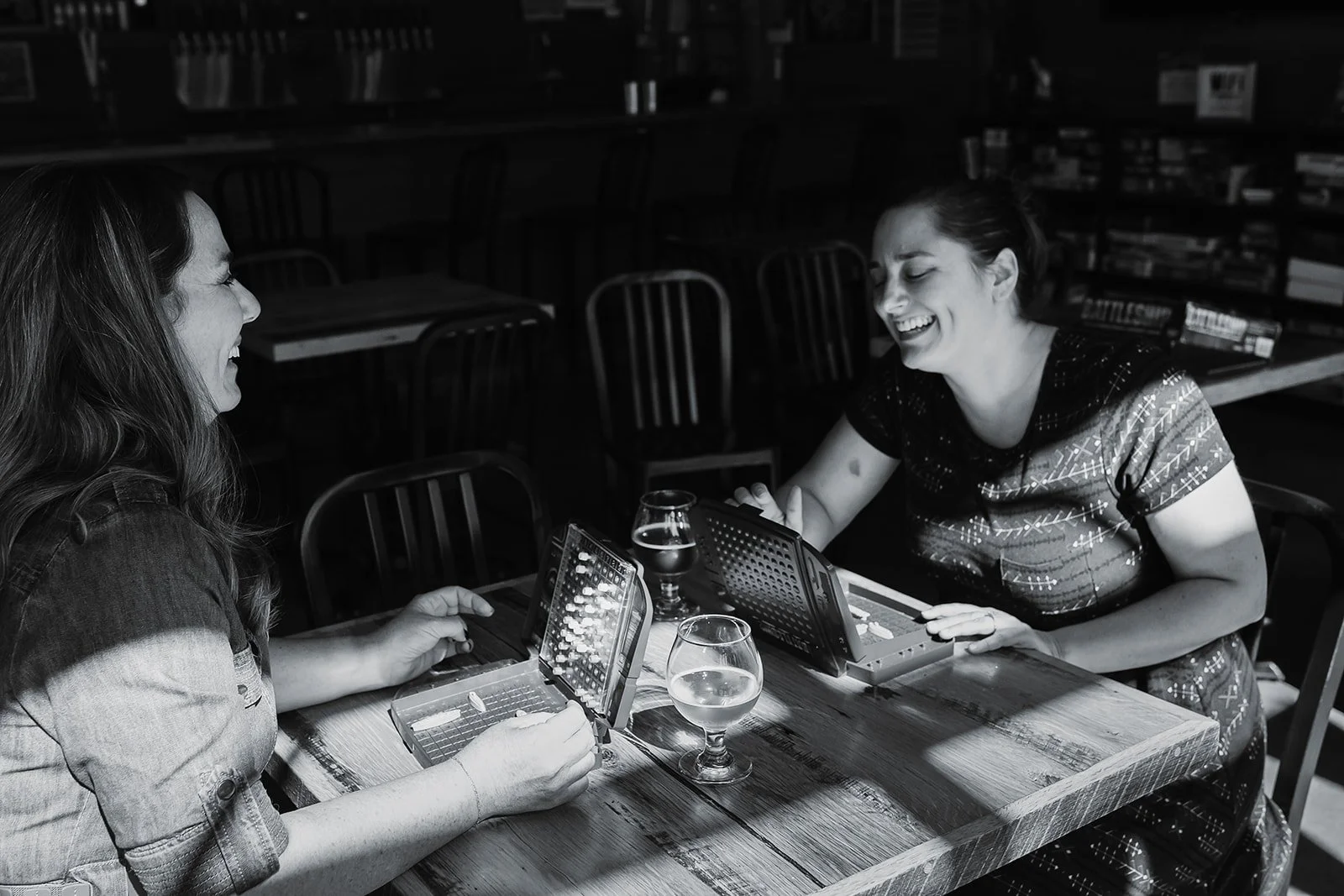 Two women sitting at a wooden table, playing a game of Connect Four, and laughing together. Two glasses of drink are on the table in front of them.