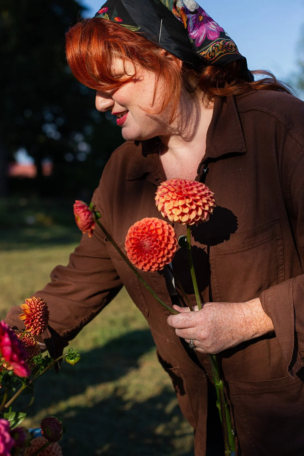 A woman with red hair and freckles holding orange and pink dahlias outdoors, wearing a dark brown jacket and a colorful headscarf, smiling while looking at the flowers.