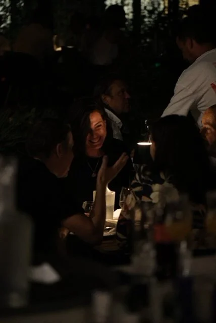 People sitting around a darkly lit table, engaged in conversation, with one woman smiling and laughing.