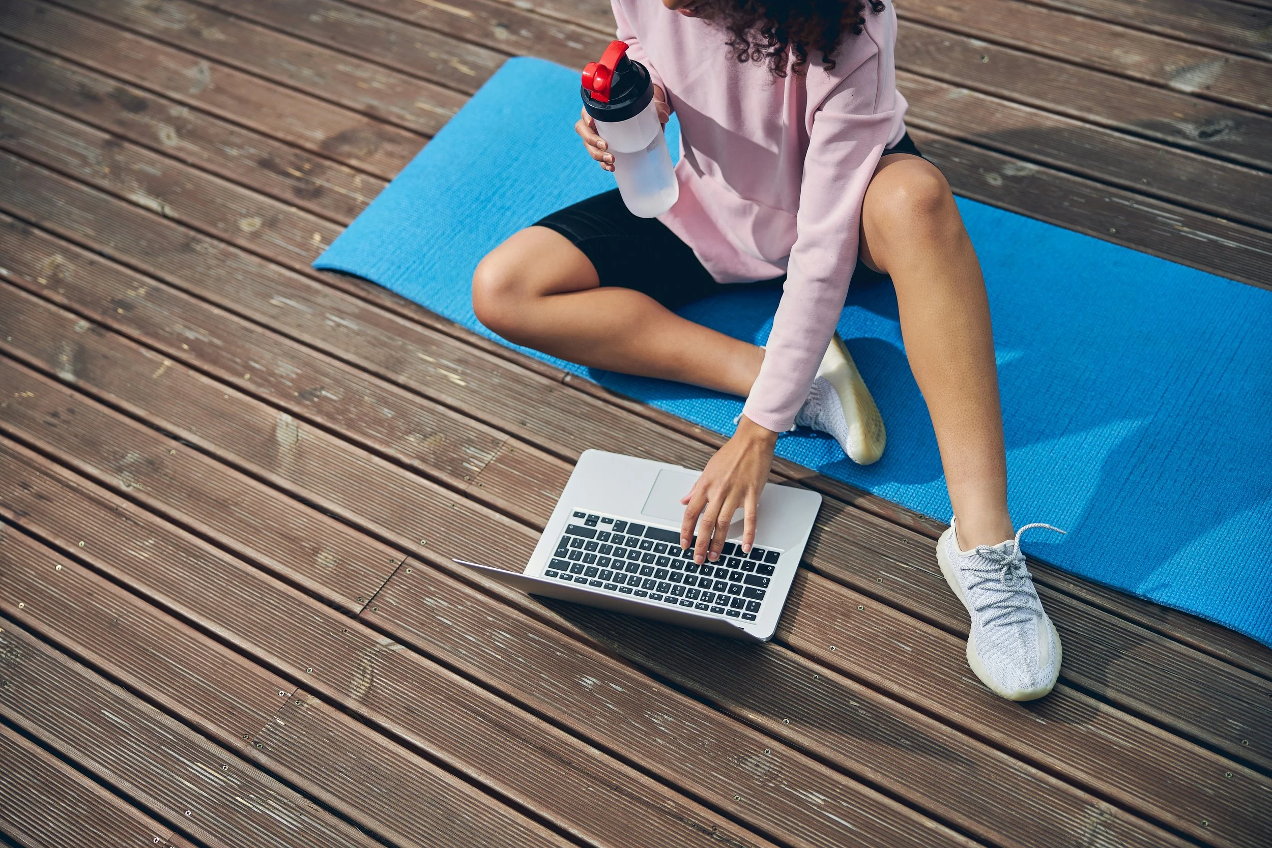 A woman sitting on a blue exercise mat on a wooden deck, holding a water bottle and typing on a laptop.