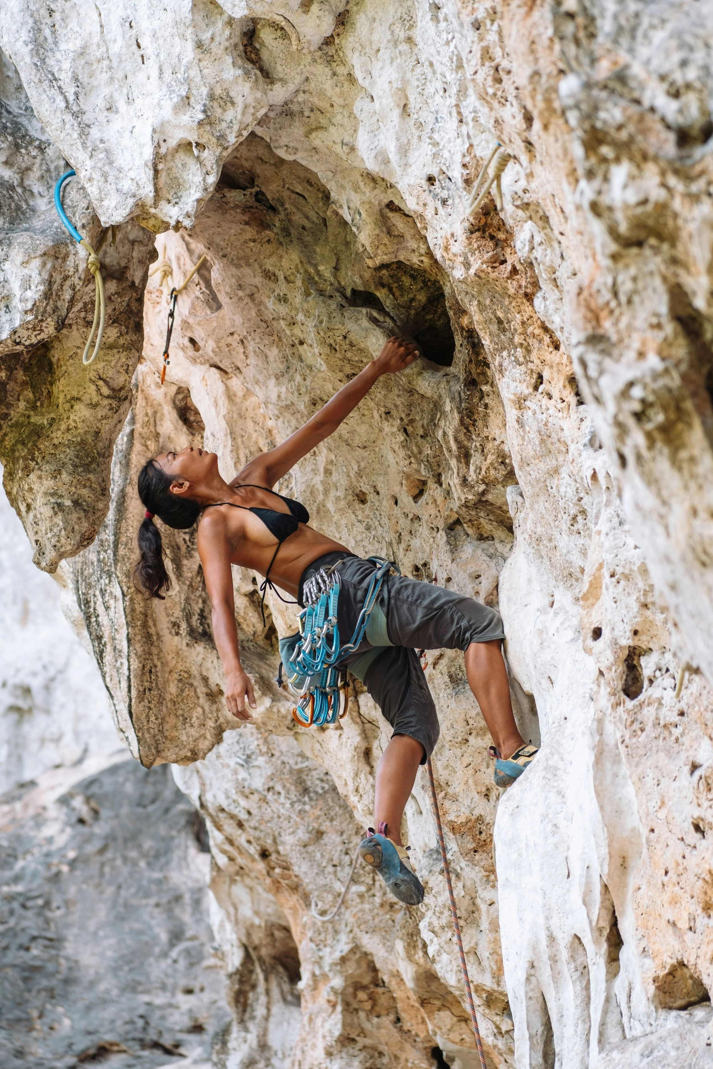 A woman climbing an overhanging limestone rock face outdoors, reaching up for a hold with a rope and quickdraws at her harness.