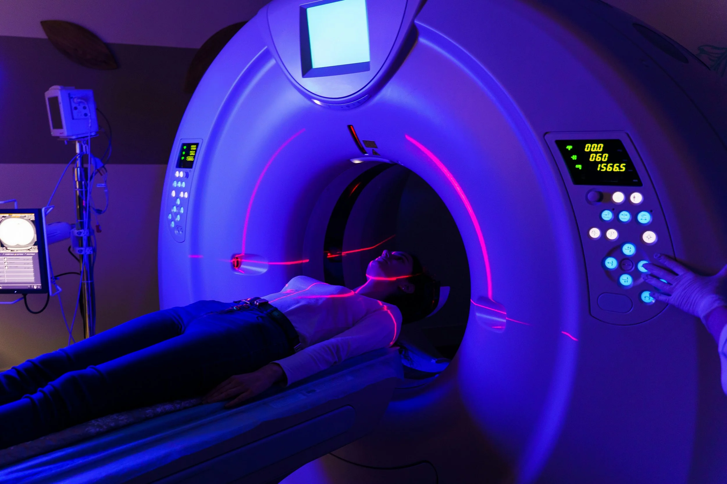 A person lying on the table of a CT scanner under blue light, with red positioning lasers across their body and a gloved hand at the control panel.