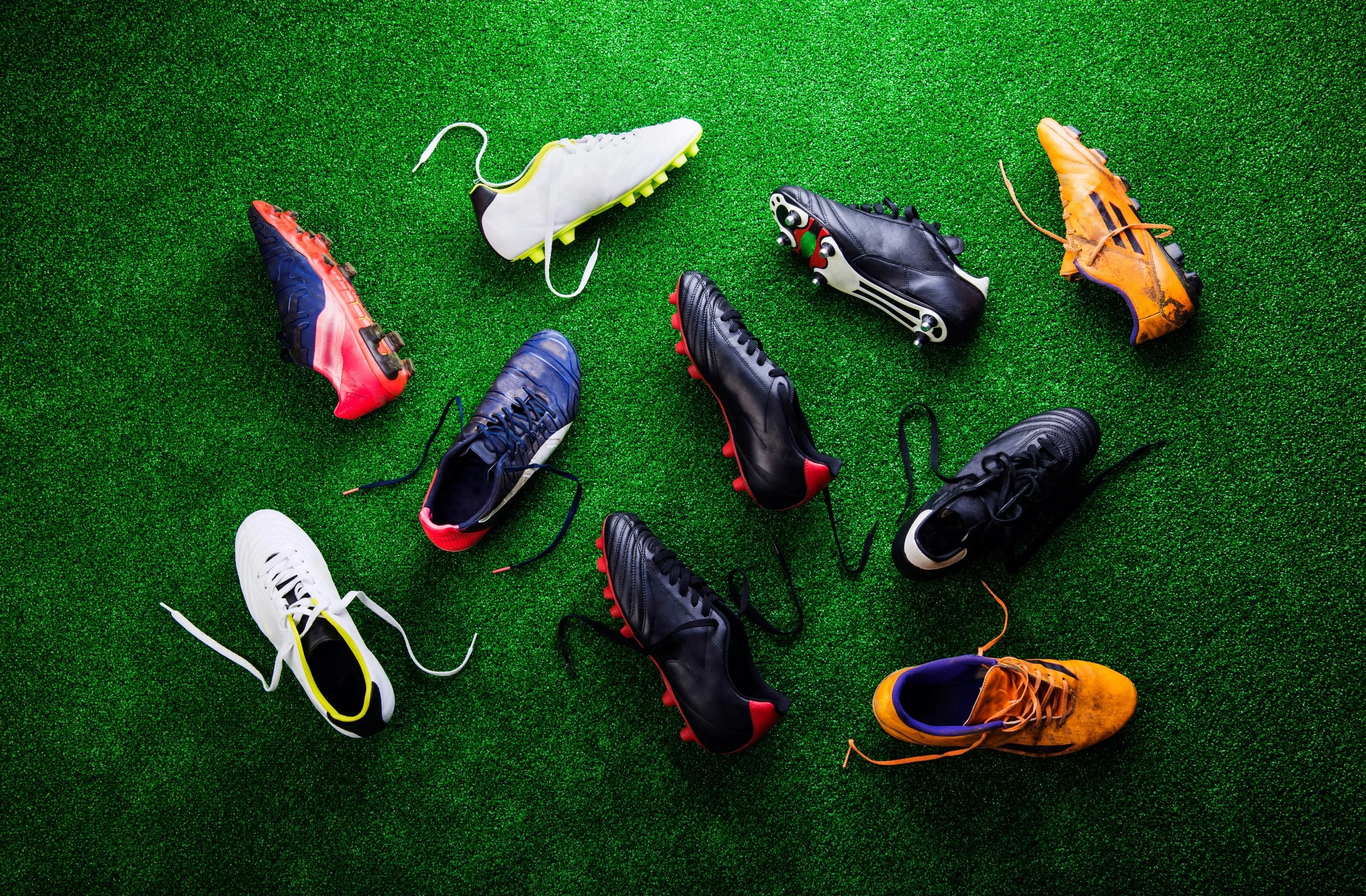 An assortment of colorful soccer cleats scattered on green artificial turf, seen from above.