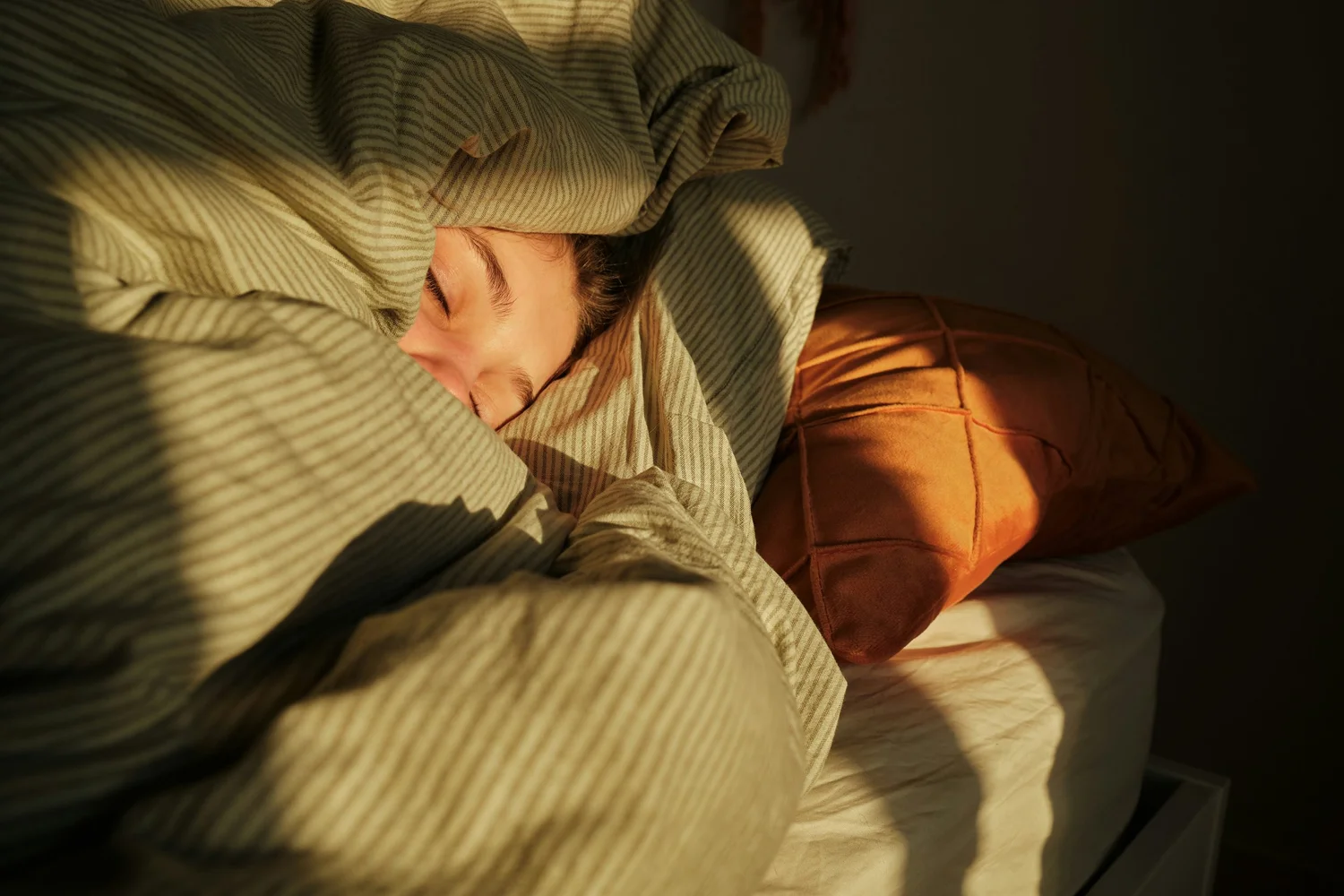 A person sleeps wrapped in a striped duvet, face lit by a stripe of warm sunlight.