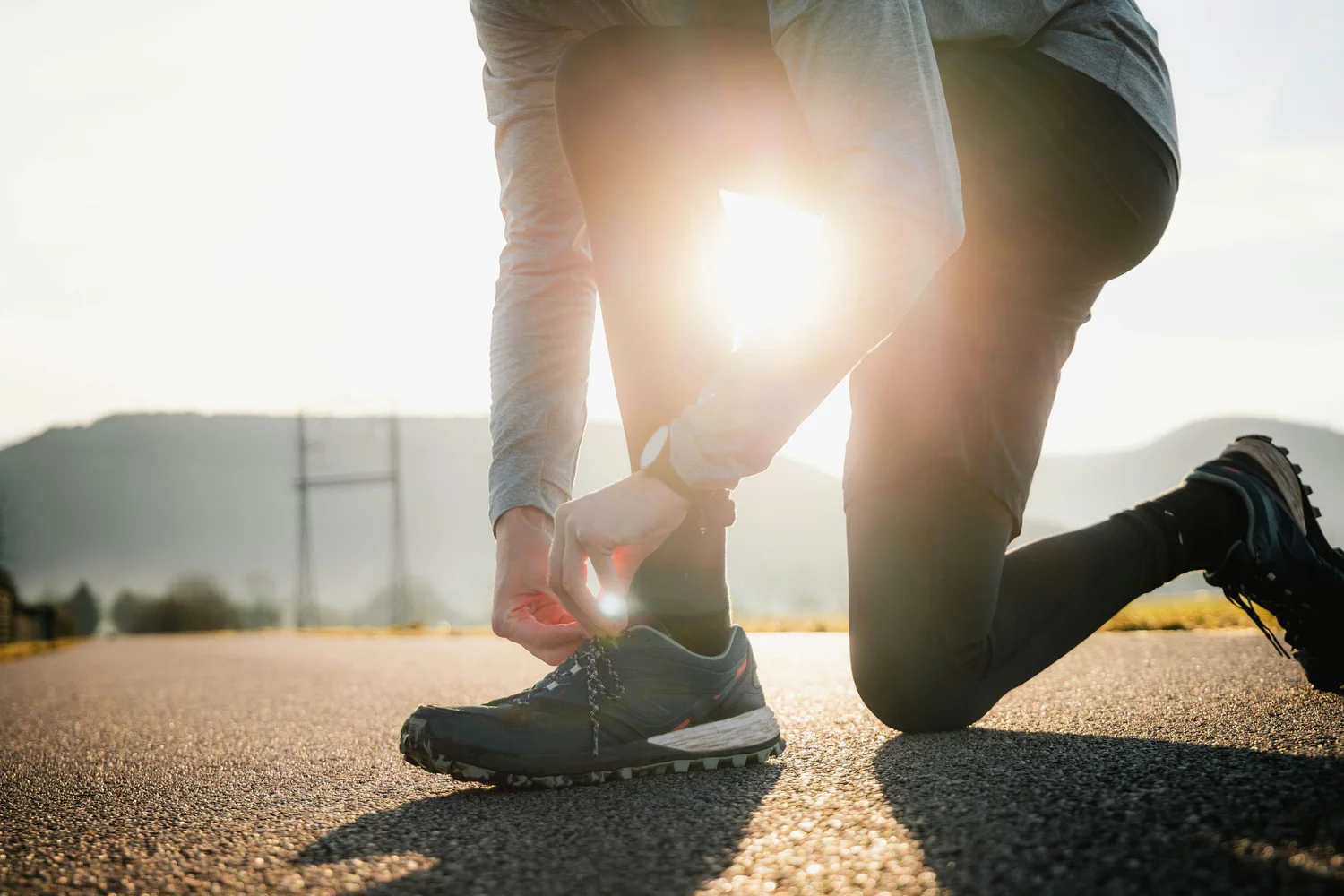 A person kneels on a road to tie their running shoe, backlit by the rising sun.
