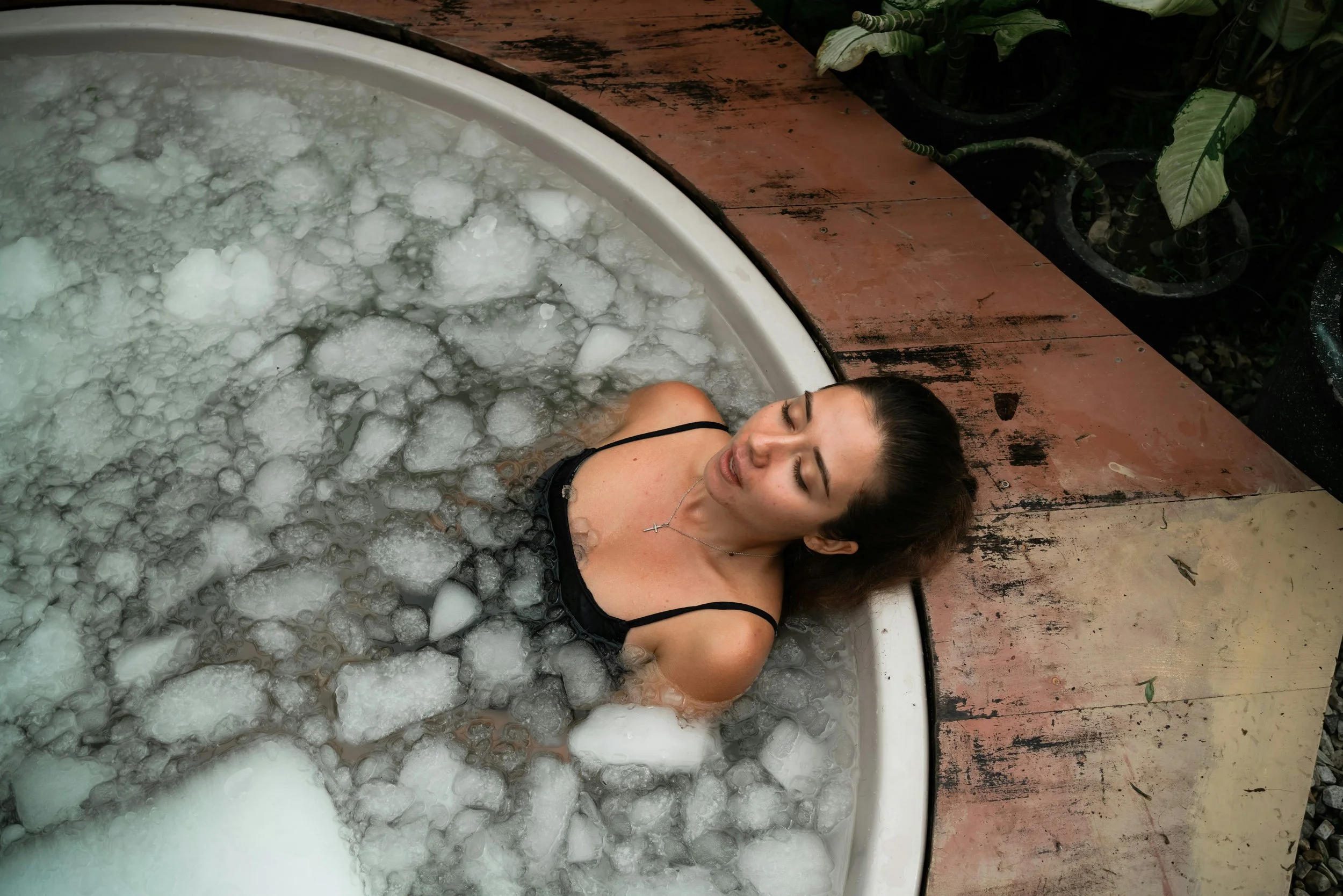 A woman with her eyes closed sitting in an outdoor tub filled with water and large chunks of ice.