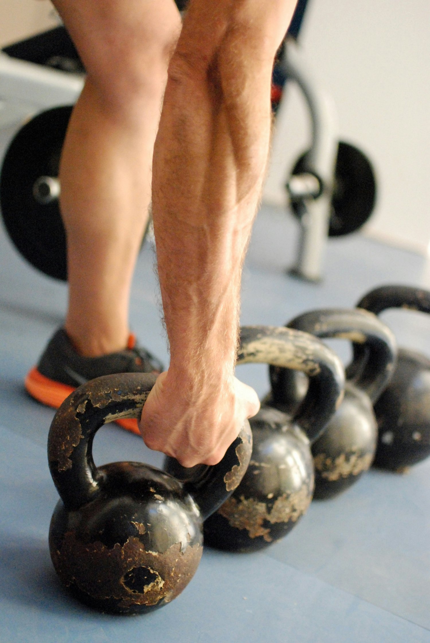 A person grips a kettlebell handle beside a row of kettlebells on a gym floor, forearm muscles visible.