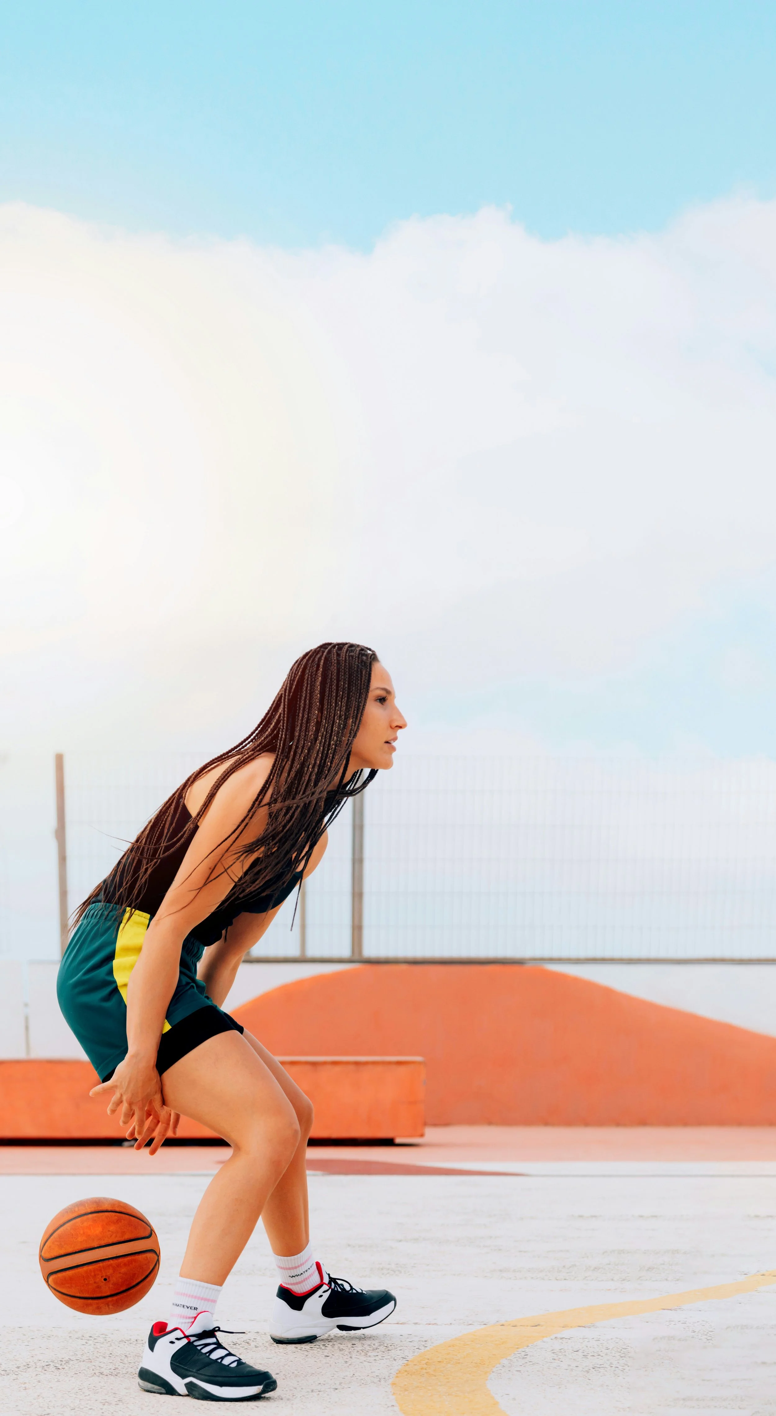 A young woman in a low athletic stance dribbling a basketball on an outdoor court under a bright sky.