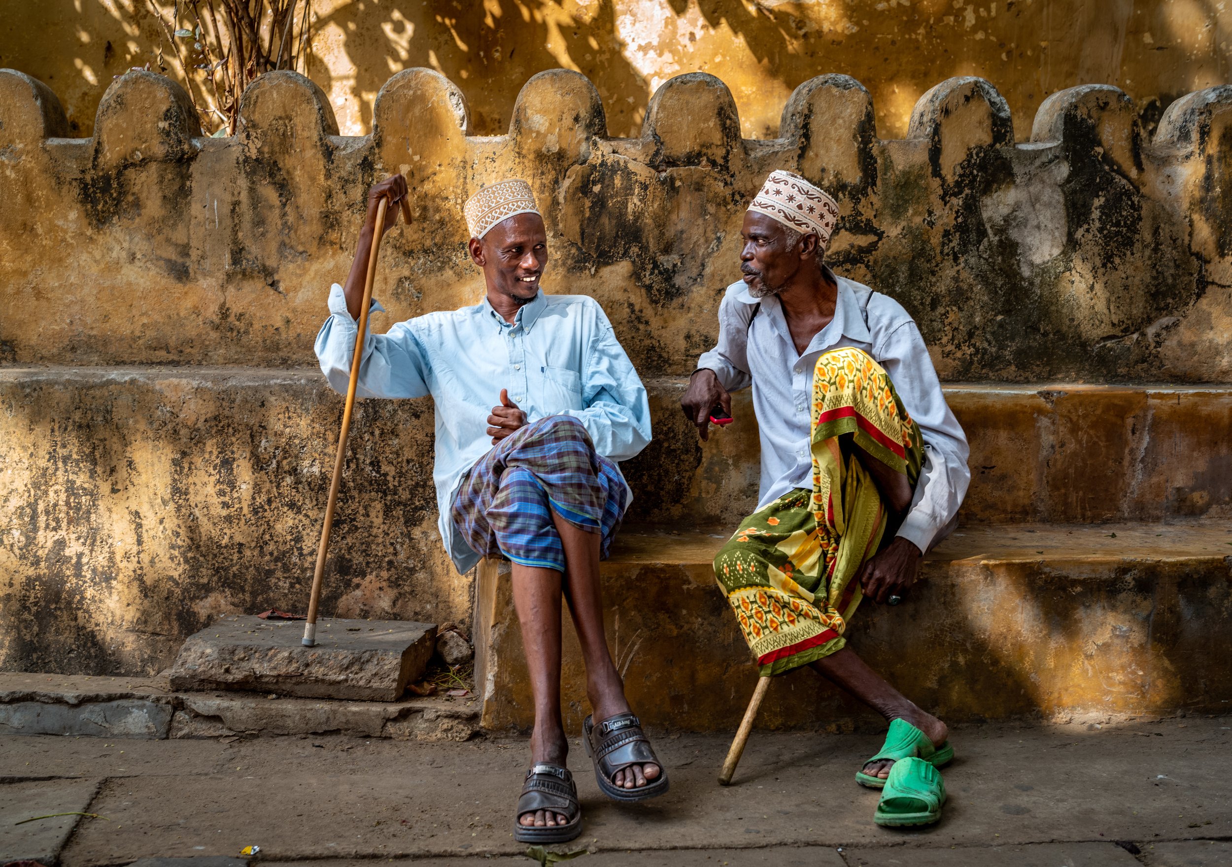 Muslim men wearing traditional koofiyad caps and colorful sarong wraps sitting on a stone bench in front of the fortress at Lamu, Kenya