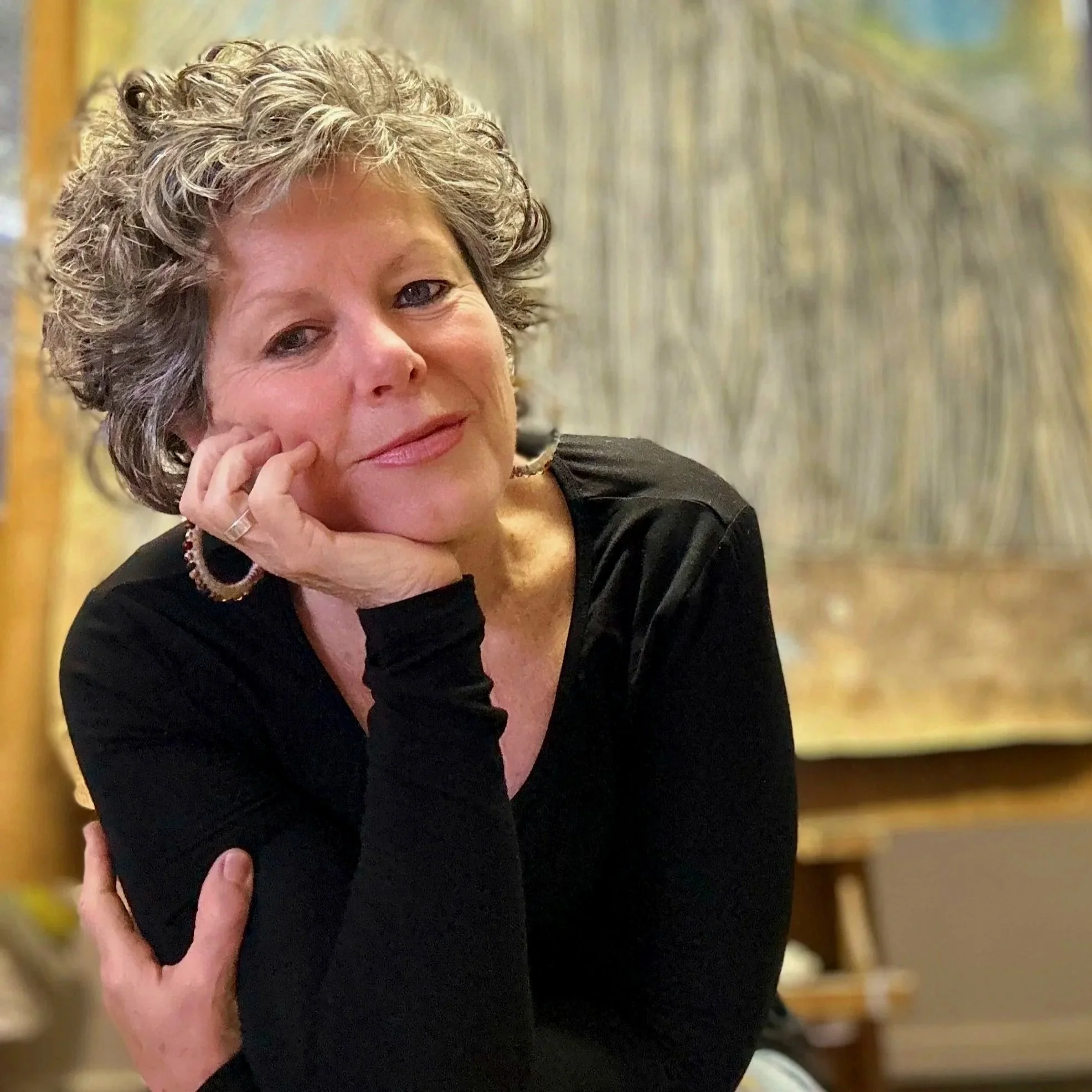 Close-up of a woman with curly gray hair leaning on her hand, smiling, wearing a black top and a bracelet, in an indoor setting with blurred background.