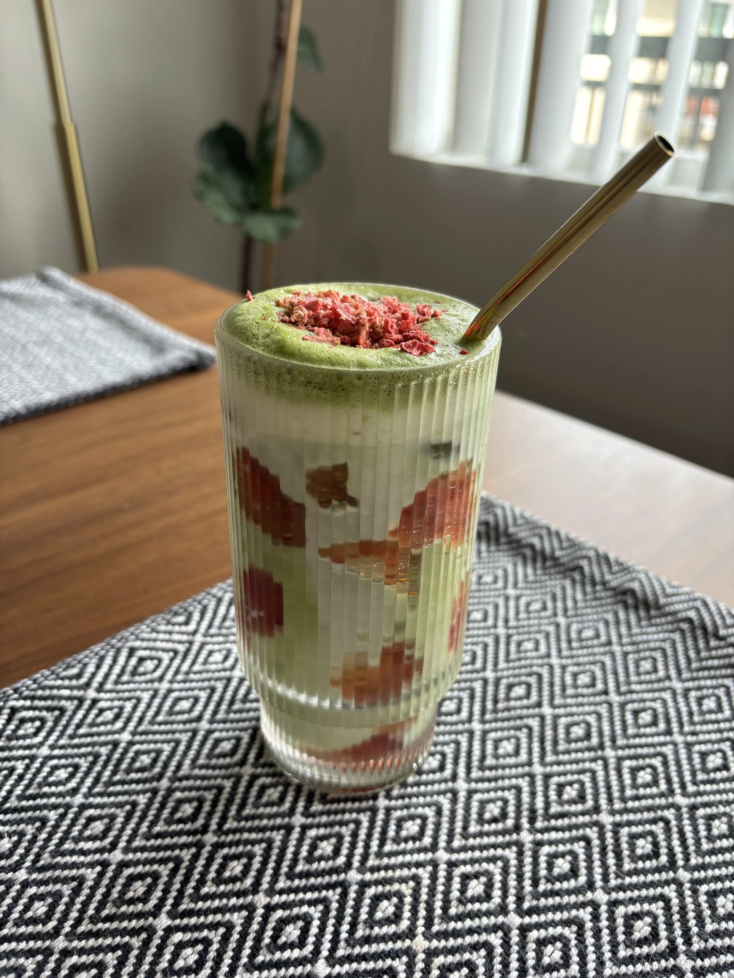 An iced matcha latte with strawberries smashed on the inside of the glass, crushed dried strawberries on top, and a gold metal straw sticking out