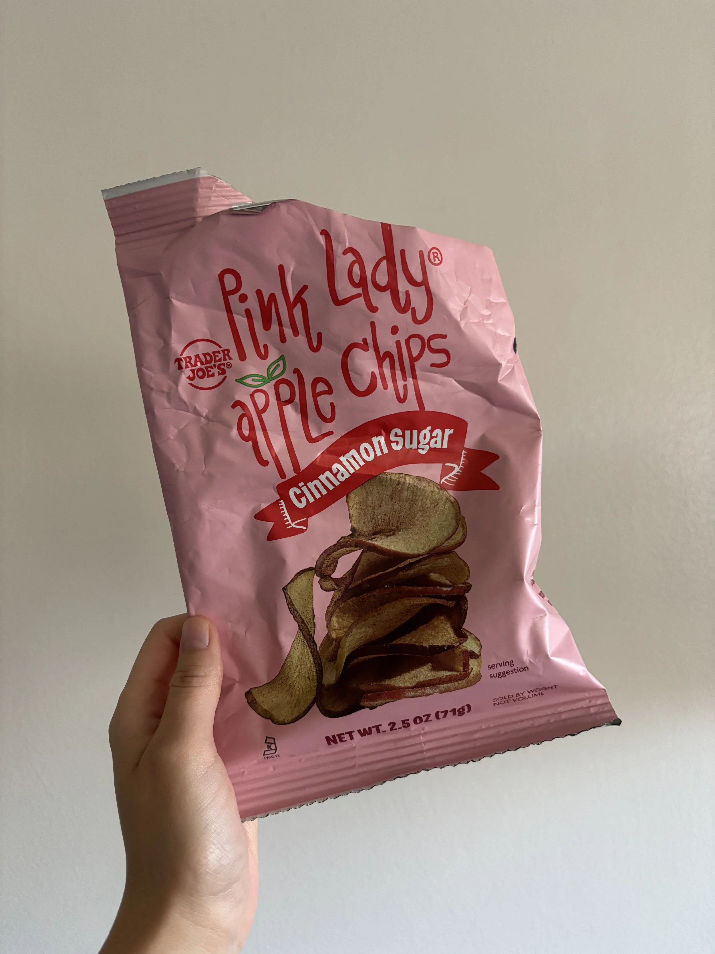 Photo of a woman's hand holding up a pink bag of Trader Joe's Pink Lady Apple Chips. The bag is opened from the top corner and has a lot of crinkles around it.