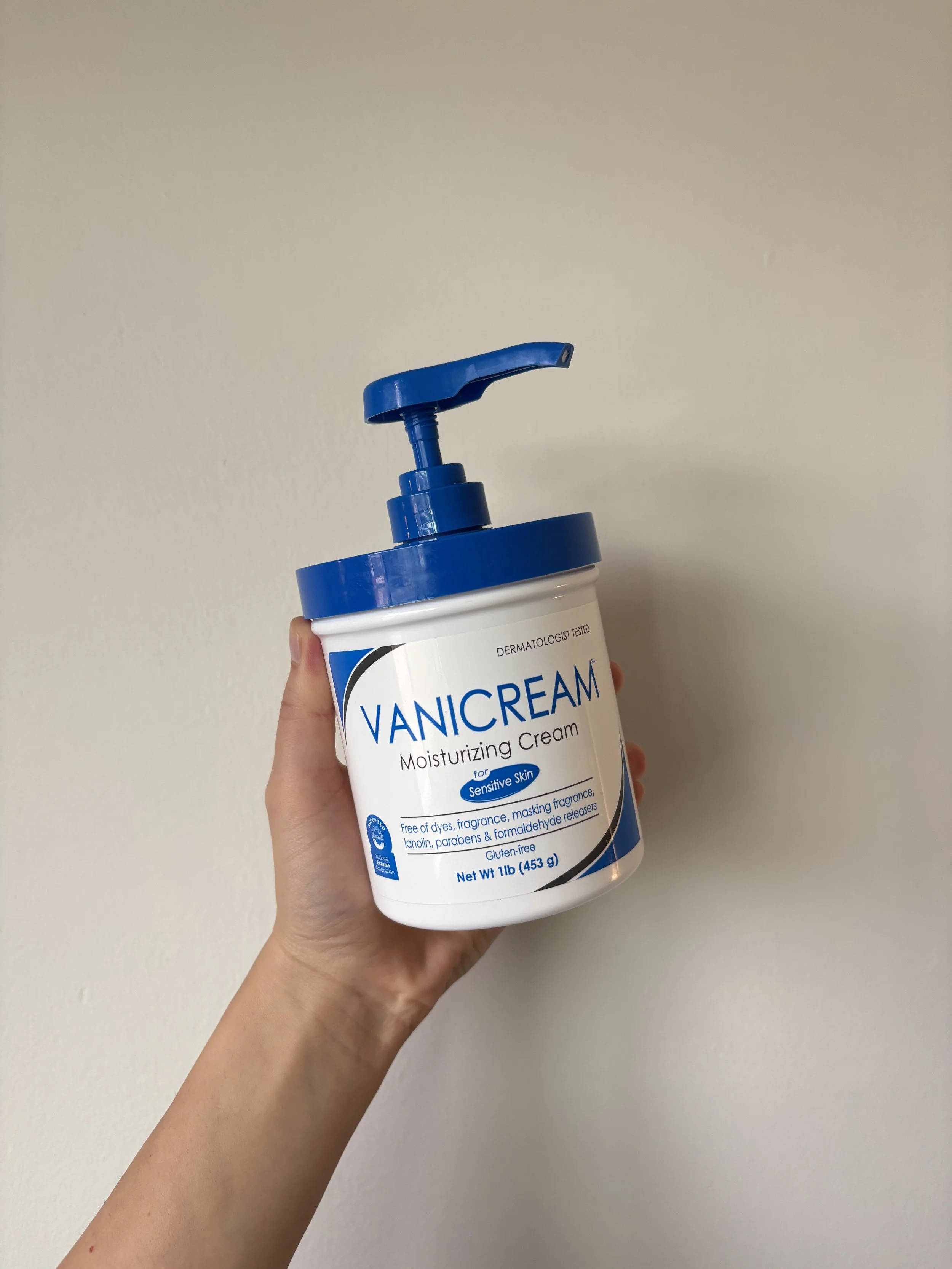 Hand holding up a Vanicream moisturizer bottle with a blue lid and pump for dispensing product
