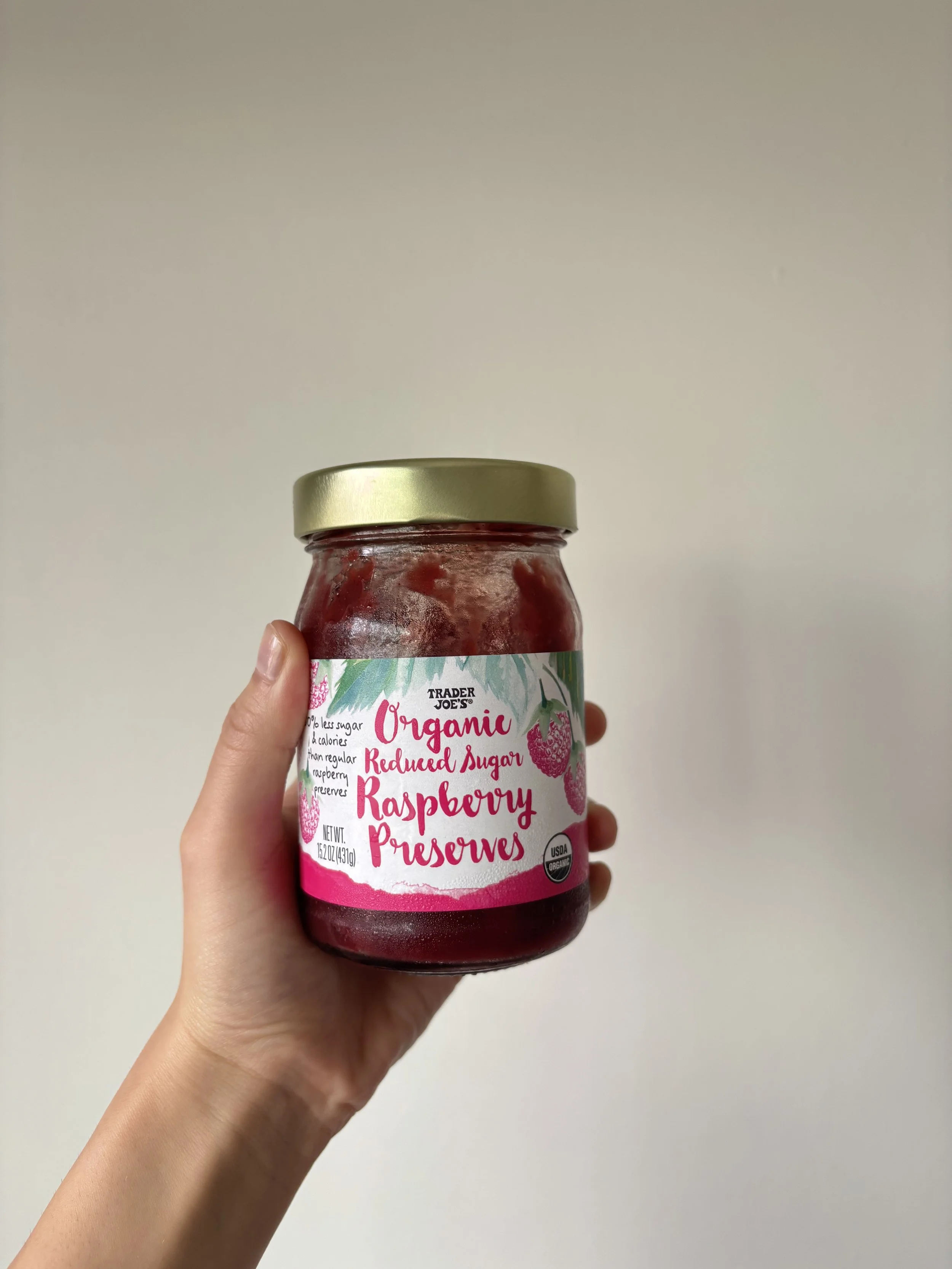 Hand holding up a small glass jar of Trader Joe's Reduced Sugar Raspberry Preserves