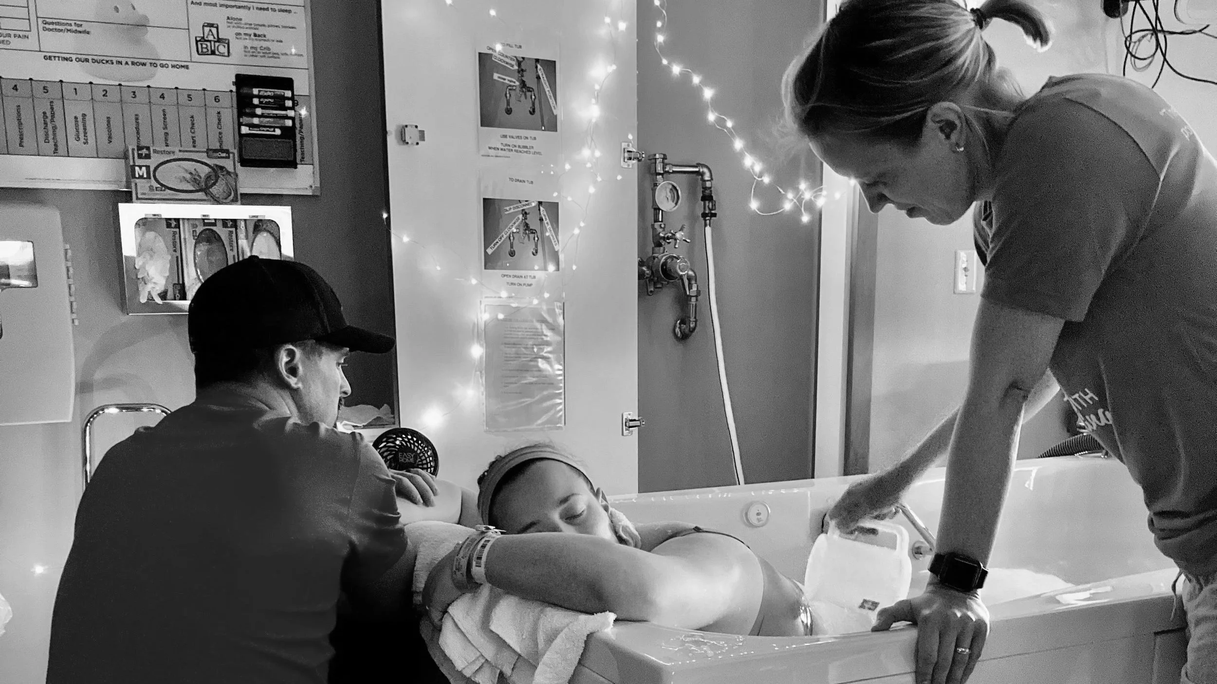 A woman in a hospital bed receiving a bath from two healthcare workers, one with a hand on her arm, in a room decorated with fairy lights and medical equipment.