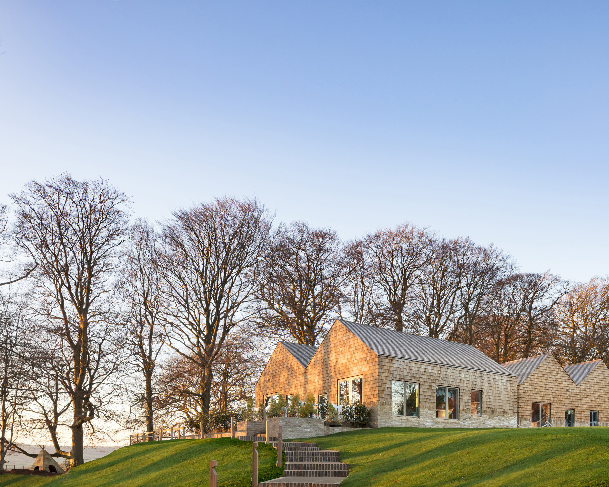 The building is conceived as a collection of timber pavilions within a woodland setting. The plan form creates separate defined rooms (pavilions) for each age stream. The plan layout provides a progression by age streams northwards through the Nursery along an active circulation space which will provide group work spaces and storage. Each of the age streams are separated by support spaces which generate a covered external play space. These relate directly to each of the rooms. The baby room is located adjacent the entrance which means that parents dropping off babies need not venture or necessarily be aware of the other spaces in the building.
