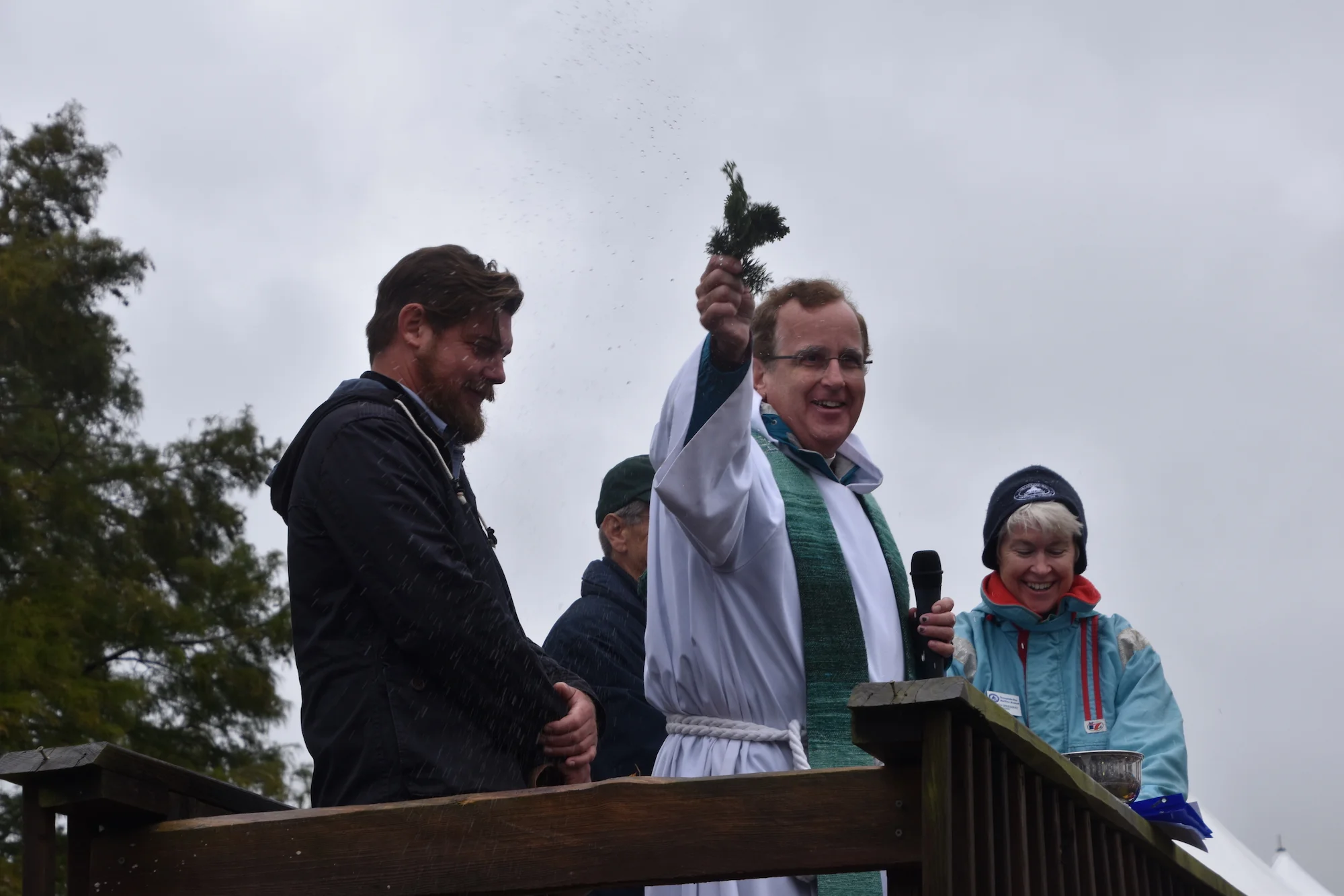 Rev. Kevin Cross from the Church of the Holy Trinity in Oxford, Md., uses holy water to bless 1889 bugeye Edna Lockwood before her return to the Miles River on Saturday, Oct. 27, 2018. Also pictured are Shipyard Manager Michael Gorman, far left, and CBMM Kristen Greenaway, right, who offered brief remarks as part of the ceremony.