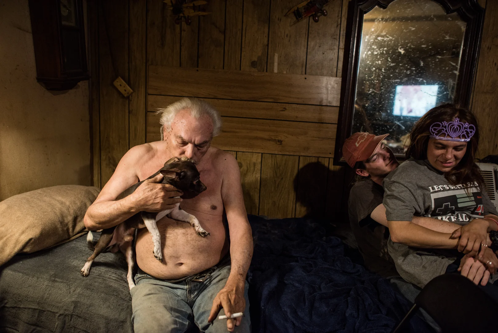 Delilah sits on Alex’s lap, as Alex’s grandfather, John, holds his dog, at John’s trailer in a remote area.