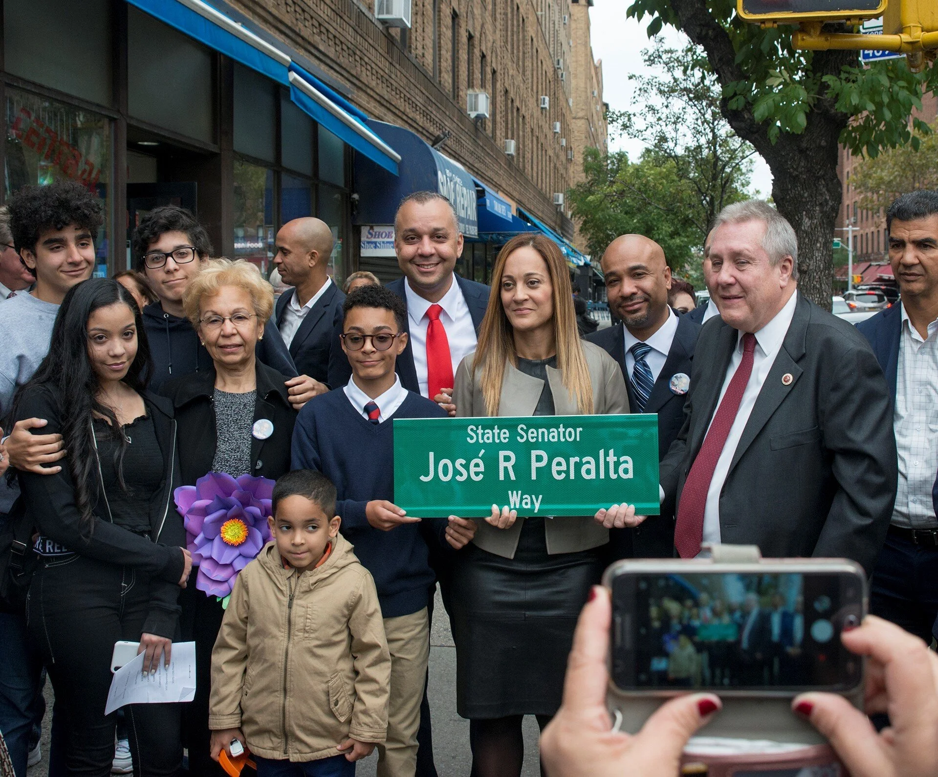 Left to right: José Peralta's surviving wife Evelyn Peralta (holding sign), his sons Matthew and Myles, other members of the Peralta family and Council Member Ydanis Rodriguez (far right) join Councilmember Daniel Dromm (next to Evelyn Peralta) at the “State Senator José R. Peralta Way” street co-naming ceremony. Photo by Jeff Reed/NYC Council.