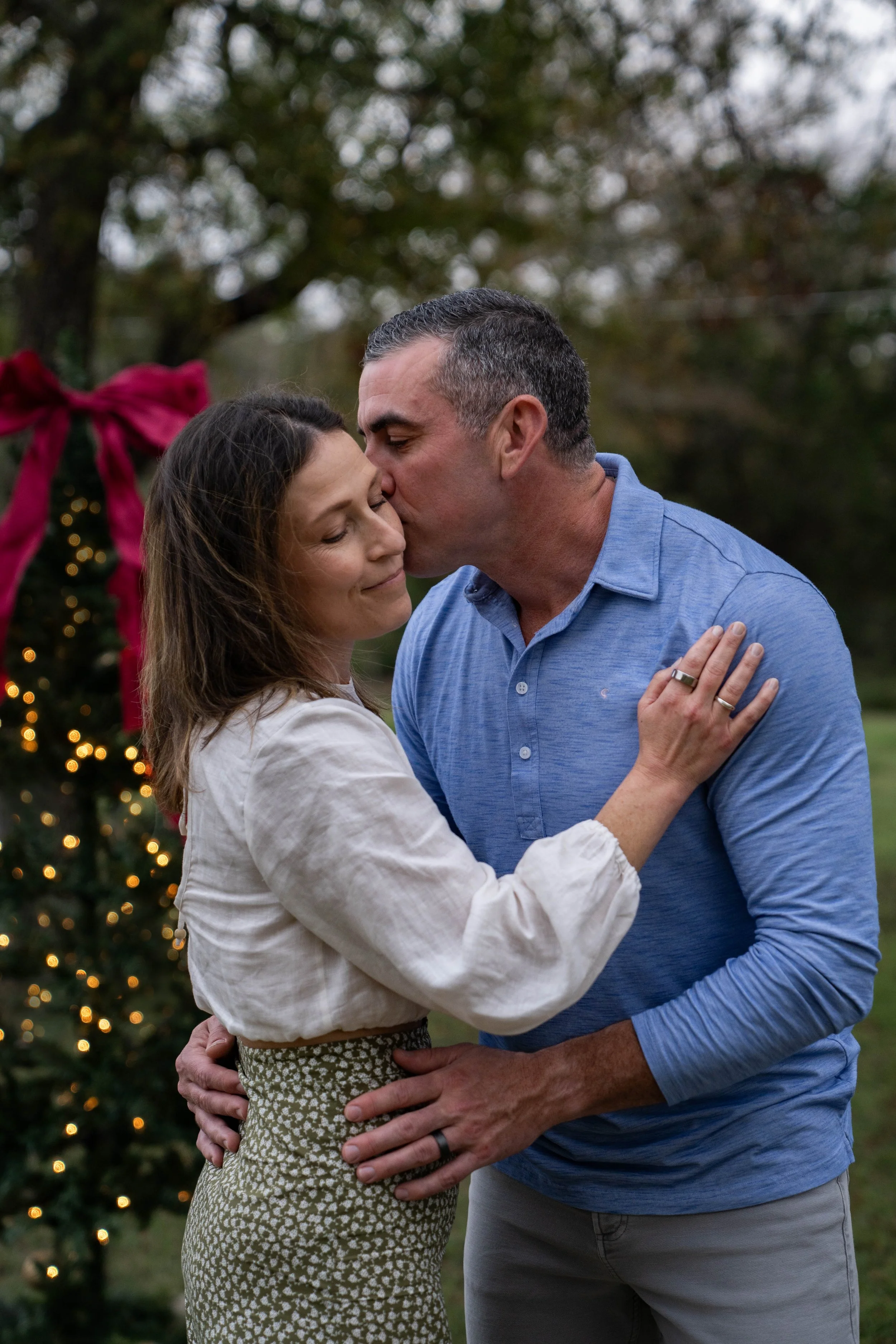 A man kissing a woman on the forehead while embracing outdoors during Christmas, with a decorated Christmas tree in the background.