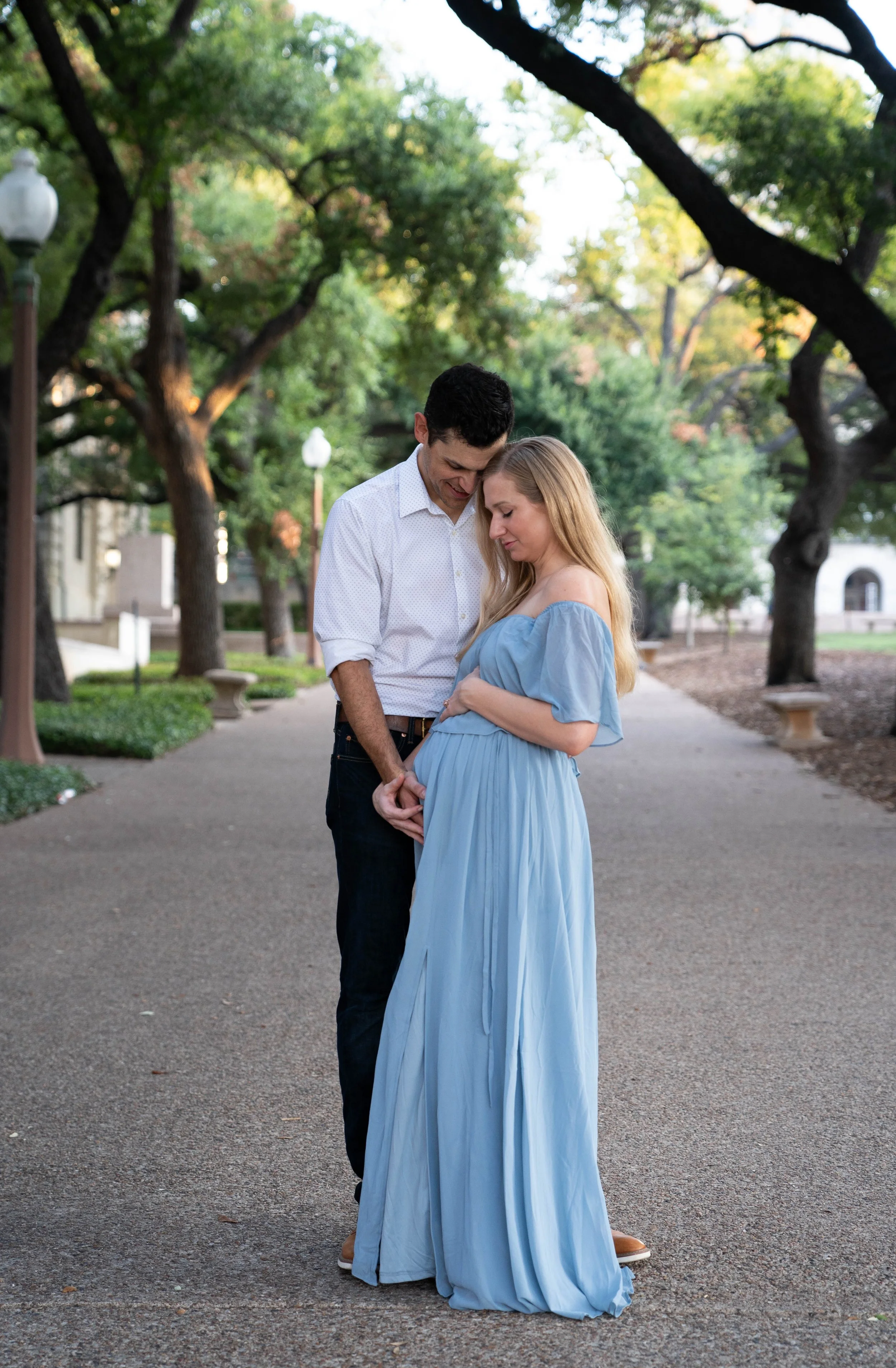 A couple standing on a park pathway, with the man gently holding the woman's pregnant belly, both looking down and smiling softly. The woman is wearing a long, light blue dress, and the man is wearing a white shirt and dark pants. Tall trees and park benches line the background.