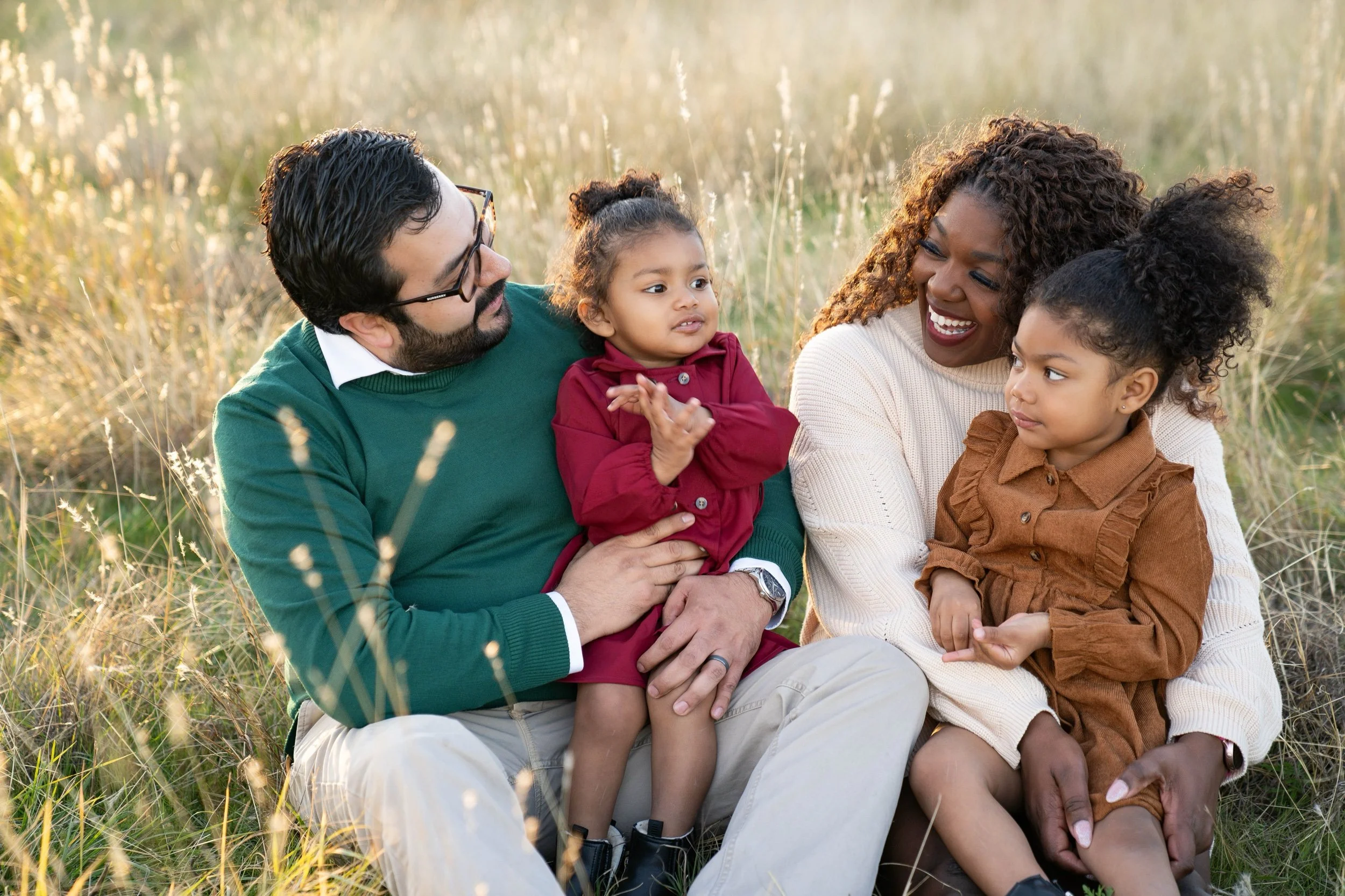 A family of four sitting in a grassy field, happily interacting. The man has glasses, dark hair, and a beard, wearing a green sweater. The woman has curly hair and a warm smile, dressed in a cream sweater. The older girl has curly hair in a ponytail,