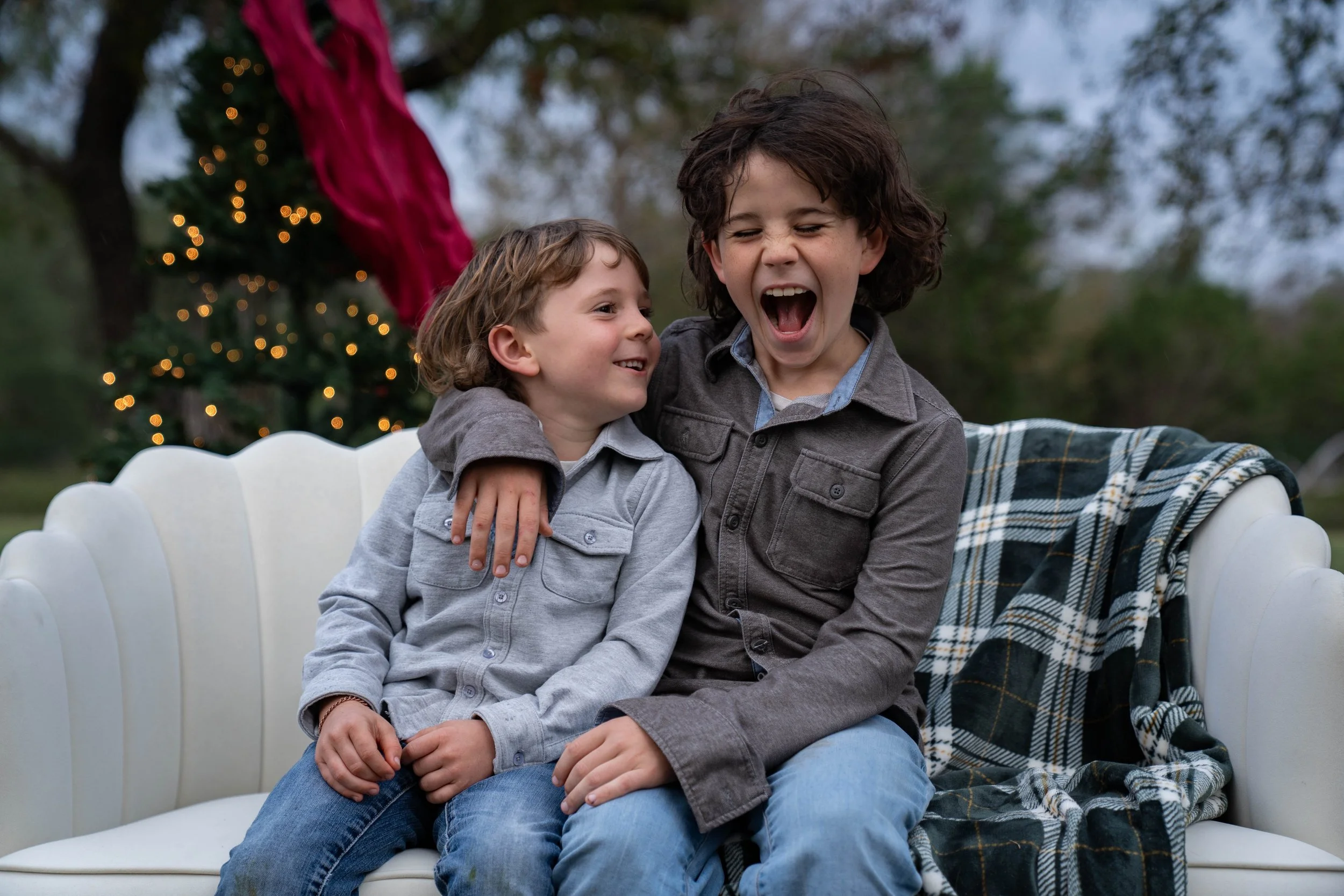 Two children, a boy and a girl, sitting on a white couch outside, smiling and laughing, with a Christmas tree in the background.