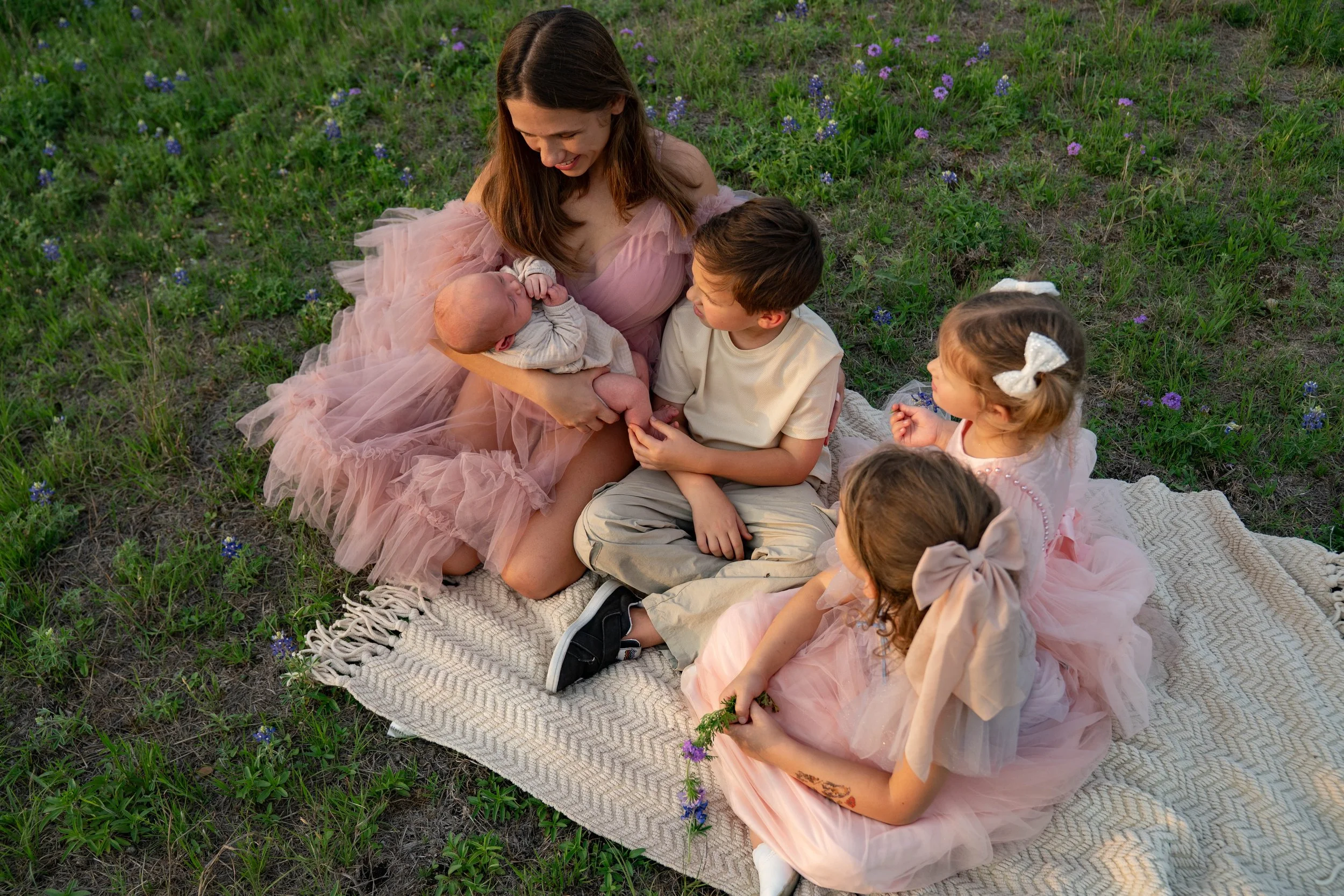 A woman, likely a mother, sitting on a patterned blanket outdoors with three children and a baby. The woman is holding the baby, which is wrapped in a light-colored blanket. The children, two girls and one boy, are sitting nearby. The girls are wearing pink dresses with bows in their hair, and one is holding a small flower. The boy is wearing a beige shirt and pants. They are all sitting on a grassy area with purple wildflowers, sharing a joyful moment together.