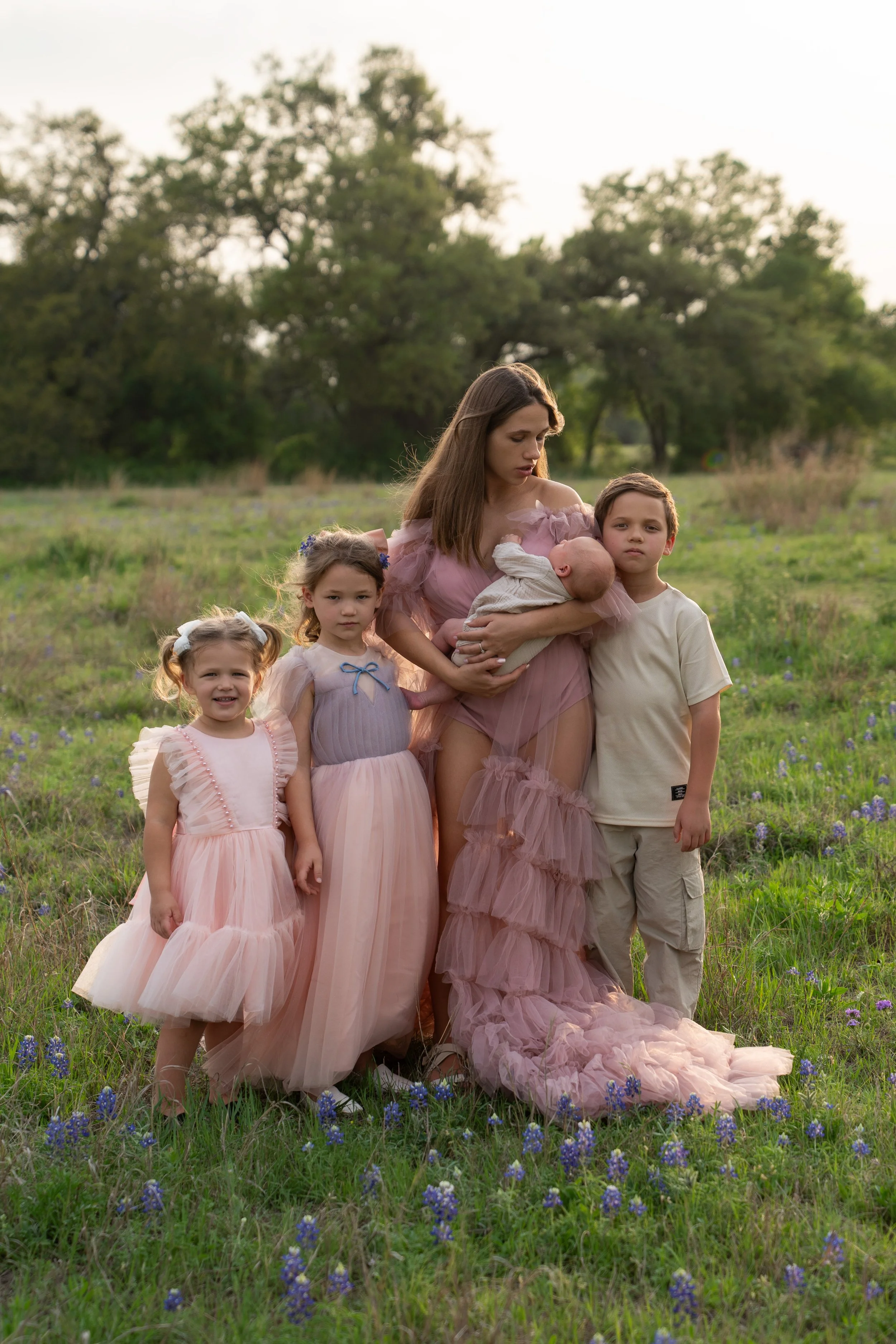 A woman holding a newborn baby in her arms, surrounded by three young children, in a green field with trees in the background.