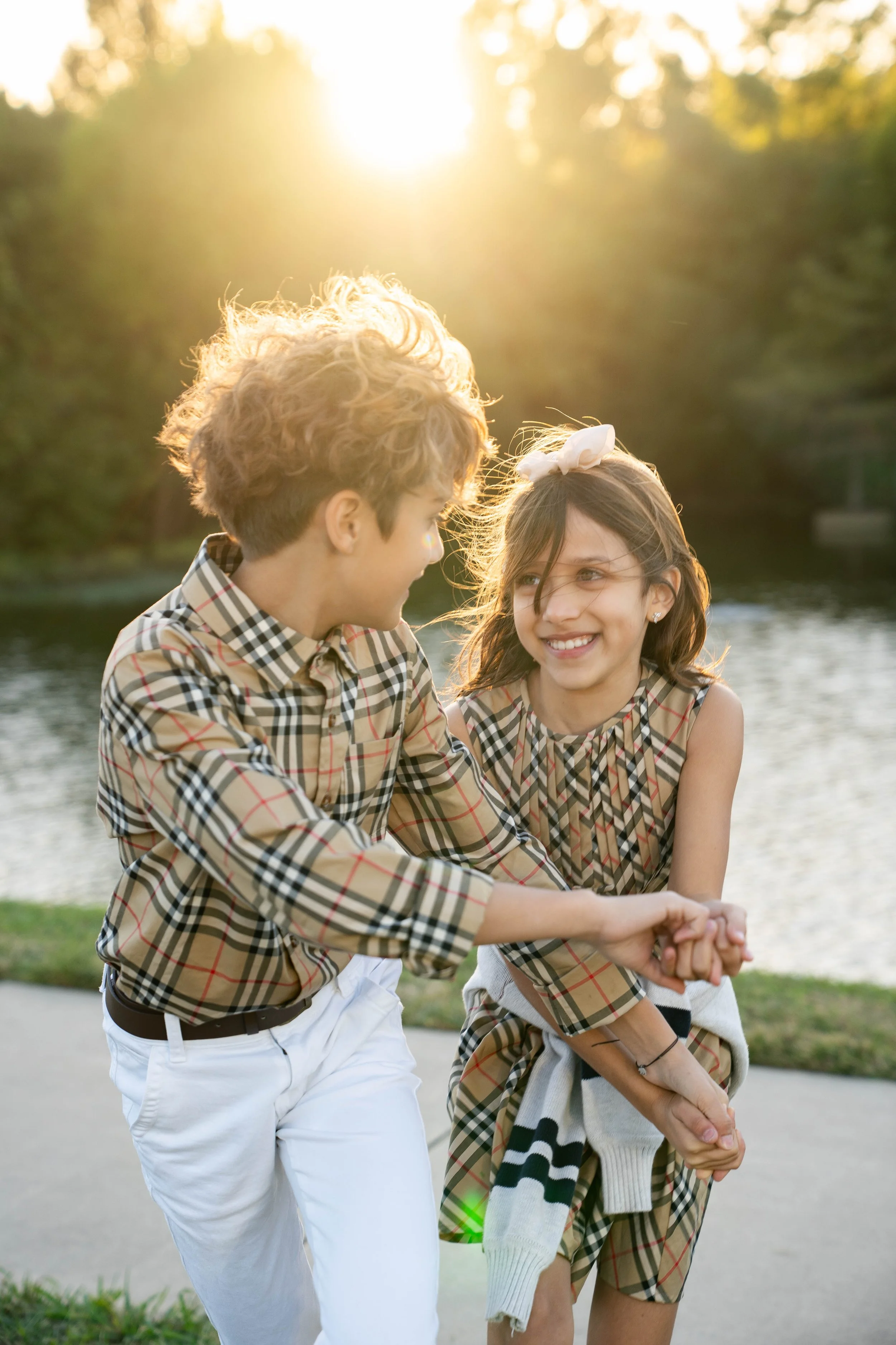 Two children, boy and girl, are outside by a body of water during sunset, smiling and holding hands.