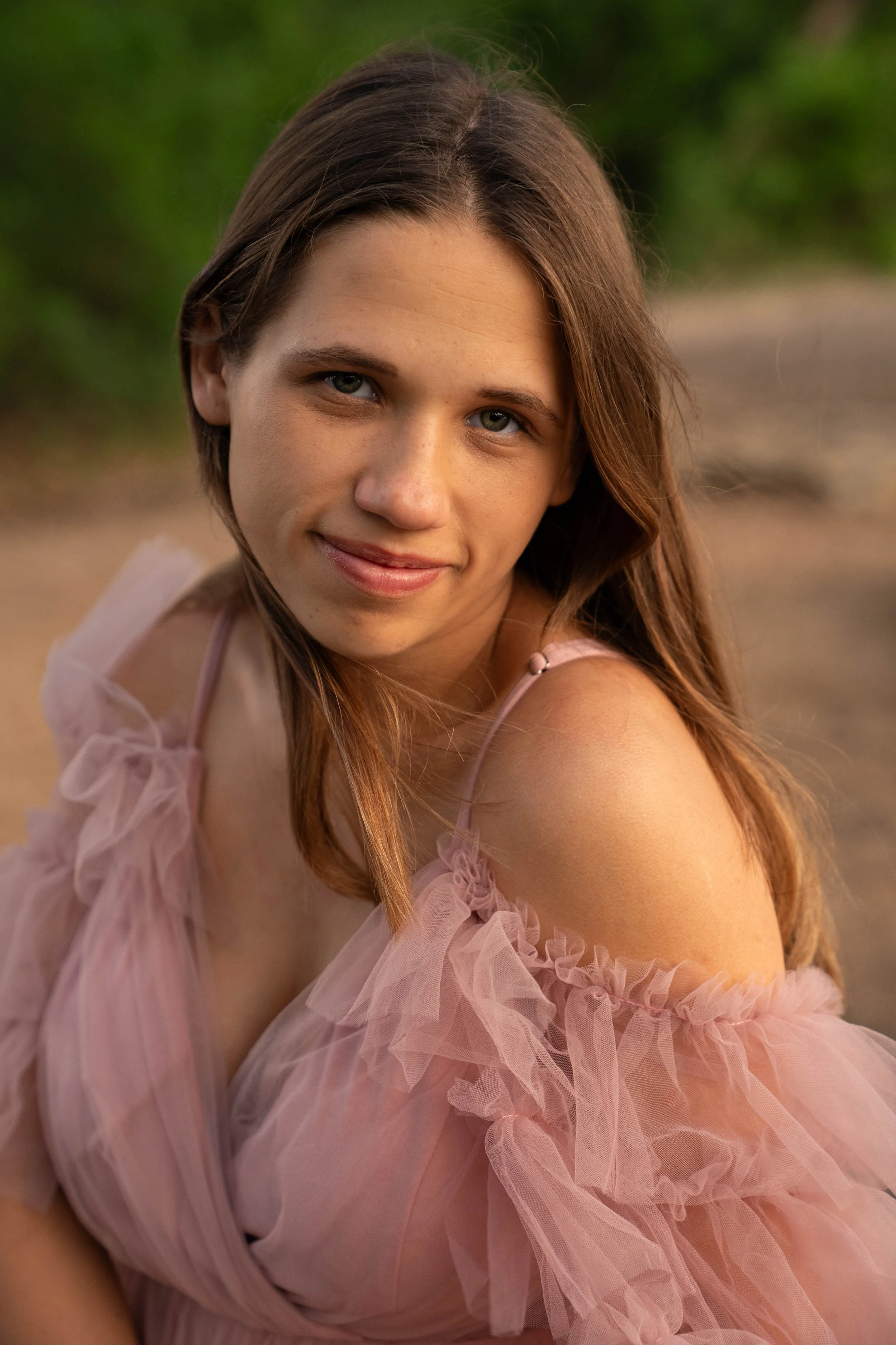 A young woman with long brown hair and fair skin smiling while looking at the camera, wearing a light pink off-shoulder ruffled dress outdoors with greenery in the background.