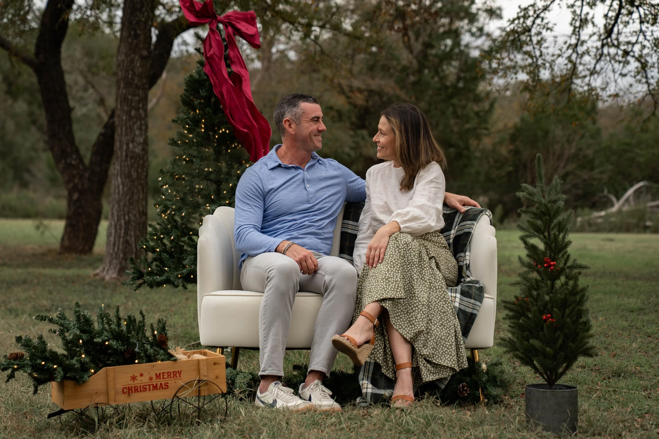 A smiling man and woman sitting together on a white sofa outdoors during Christmas, surrounded by small decorated Christmas trees and a festive wooden crate, with a red ribbon and green trees in the background.