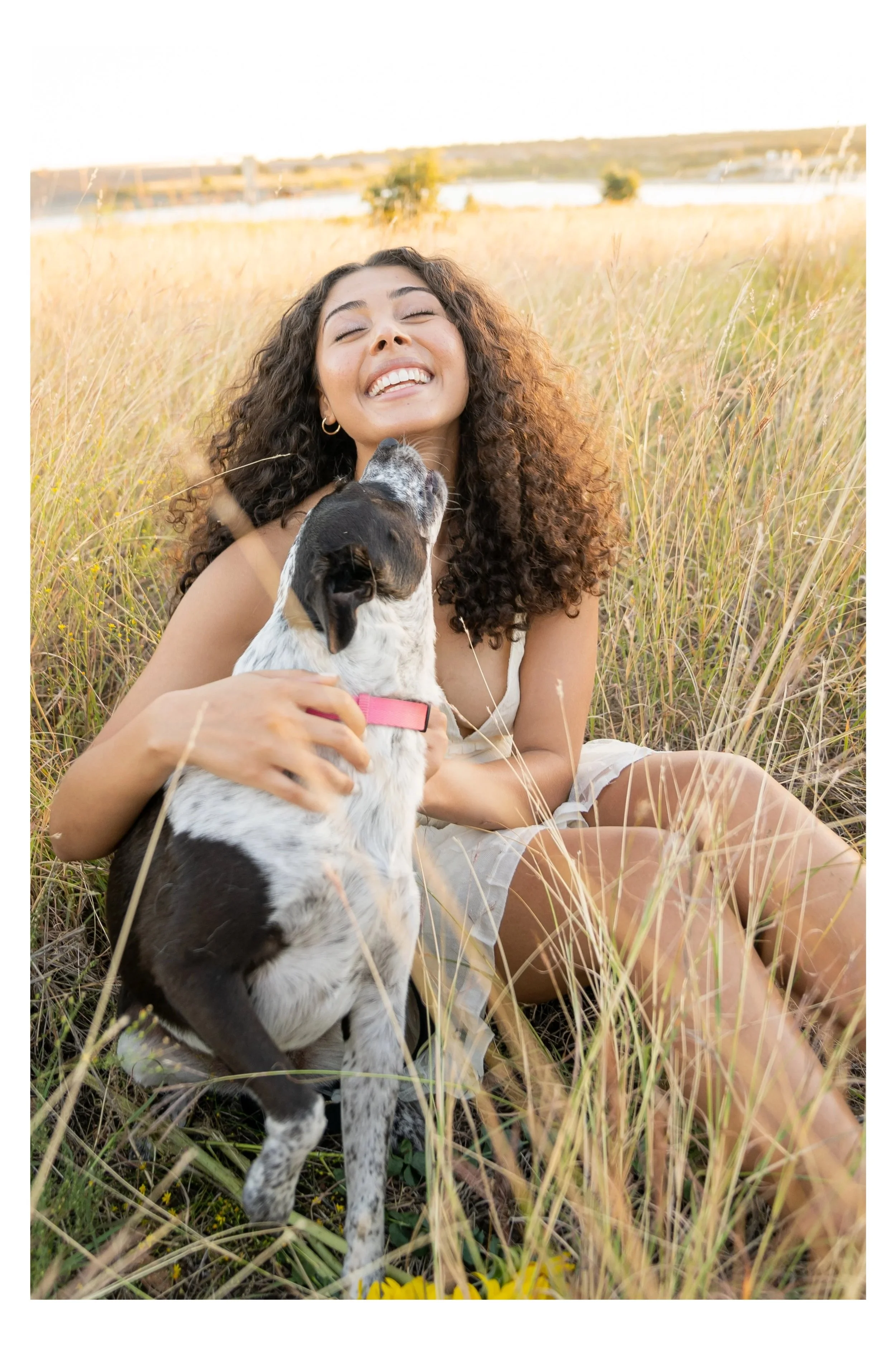 A young woman with curly hair smiling and holding a puppy in a grassy field during sunset.