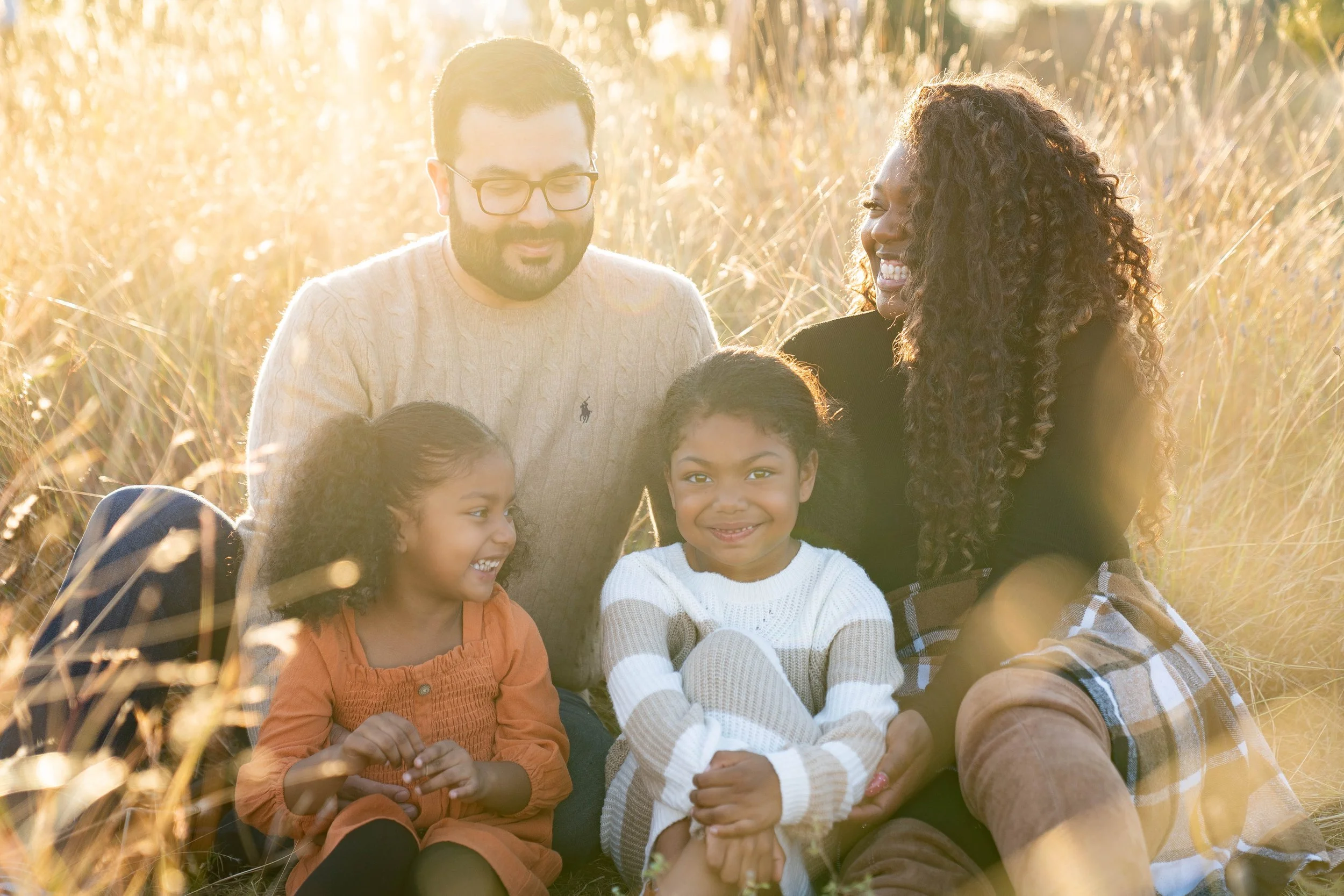 A family of four sitting on the grass in a field during golden hour, smiling and enjoying each other's company.