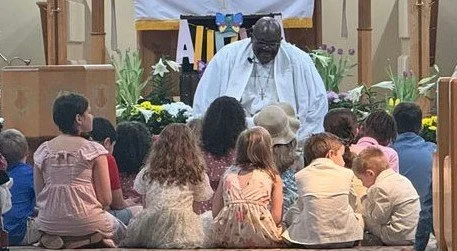 Man speaking in front of a group of children seated on the floor at an outdoor event with plants and wooden structure in the background.
