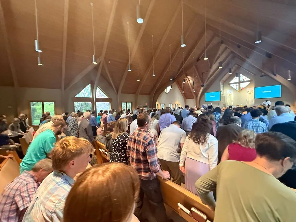 A large congregation of people praying or singing in a church with wooden ceiling and windows.