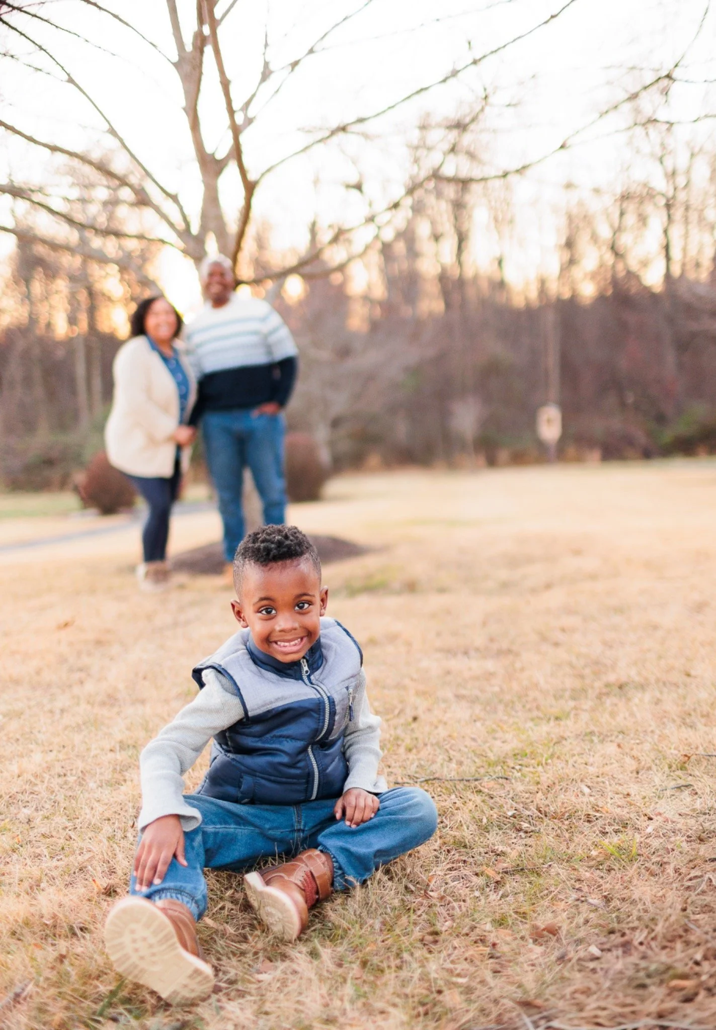 Young boy smiling while sitting on grass with his parents blurred in the background at the Shrine of St. Anthony in Clarksville, Maryland.