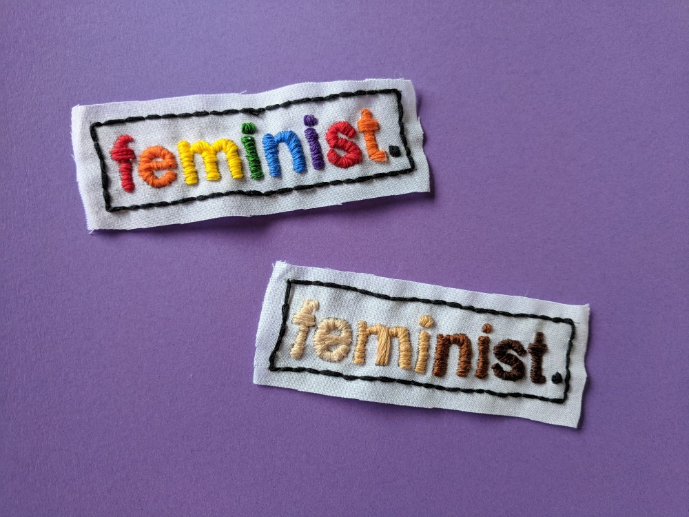 Image of two rectangle patches with one in rainbow text, “feminist” and multi-nude coloured text, “feminist”