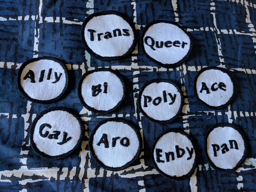 Image of white circle patches with separate text, “trans, queer, ally, bi, poly, ace, gay, aro, enby, pan”