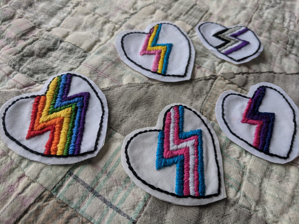 Image of heart-shaped patches with a pride flag lightning bolt in the middle of each heart - pride, trans, pansexual, asexual, and bisexual flag are shown.