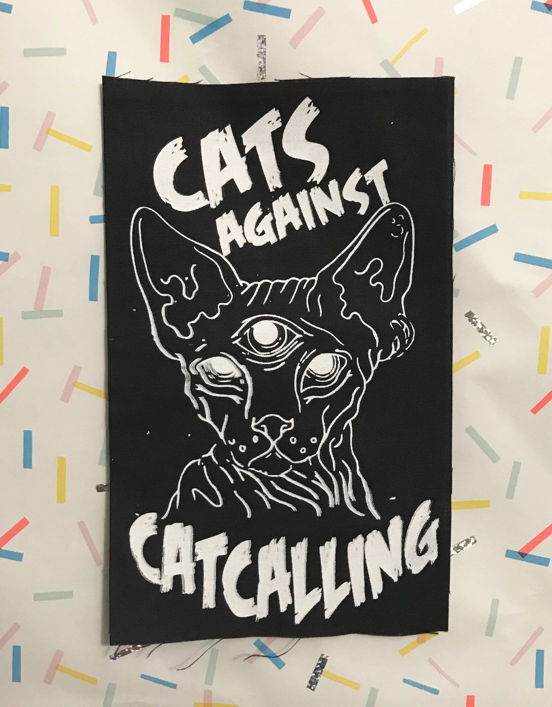 Image of black patch with white three-eyed sphynx cat and text, “cats against catcalling”